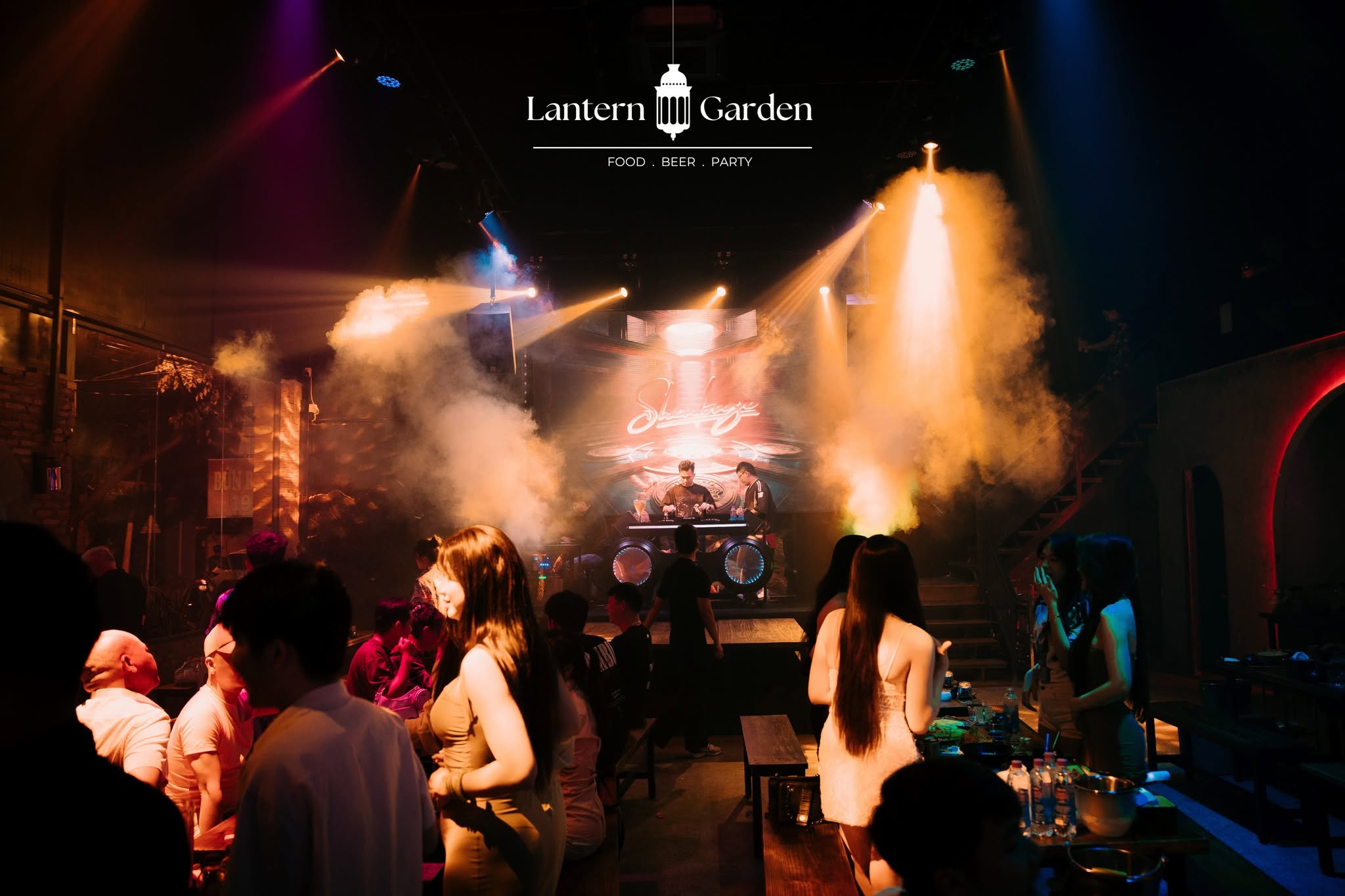 A dimly lit nightclub or event space with a band performing on stage under dramatic colored lights and smoke. Several people are standing and dancing in the foreground, and a logo reading 'Lantern Garden' is visible at the top center.