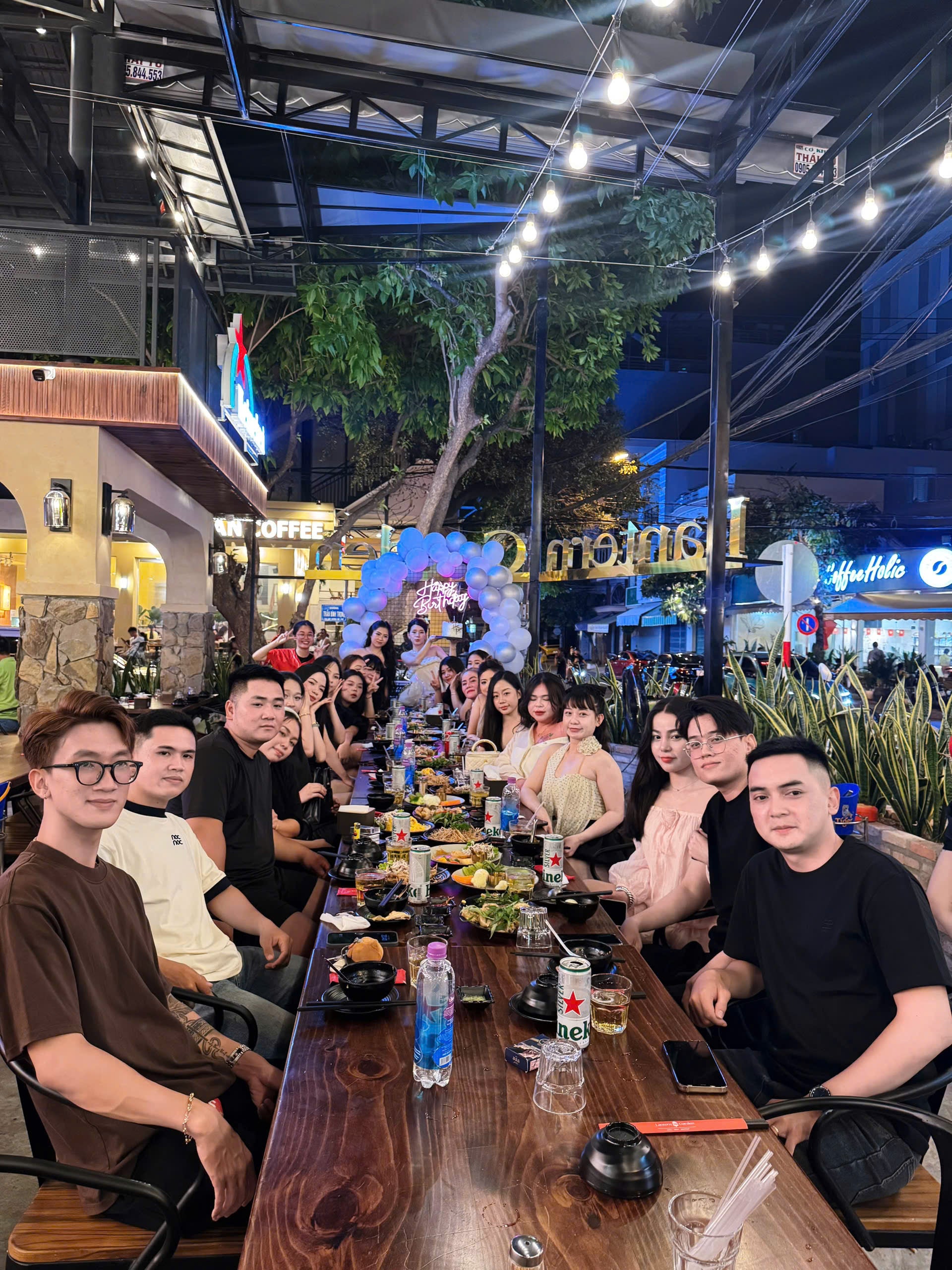 Group of people sitting around a long table at night in an outdoor setting with lights and buildings in the background.