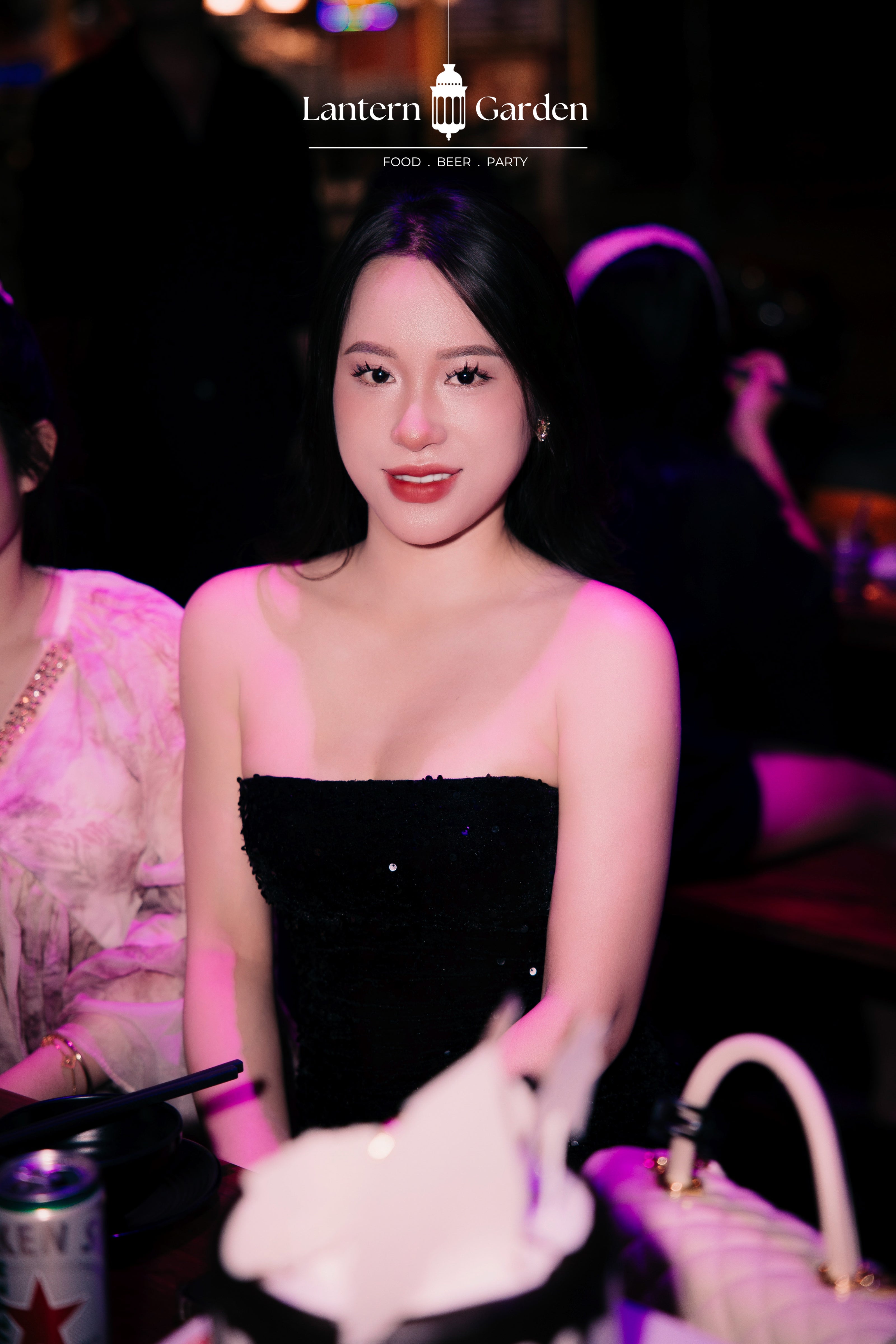A woman wearing a strapless black sequined dress at a dimly lit event or club, with pink ambient lighting and a visible 'Lantern Garden' logo at the top of the image.