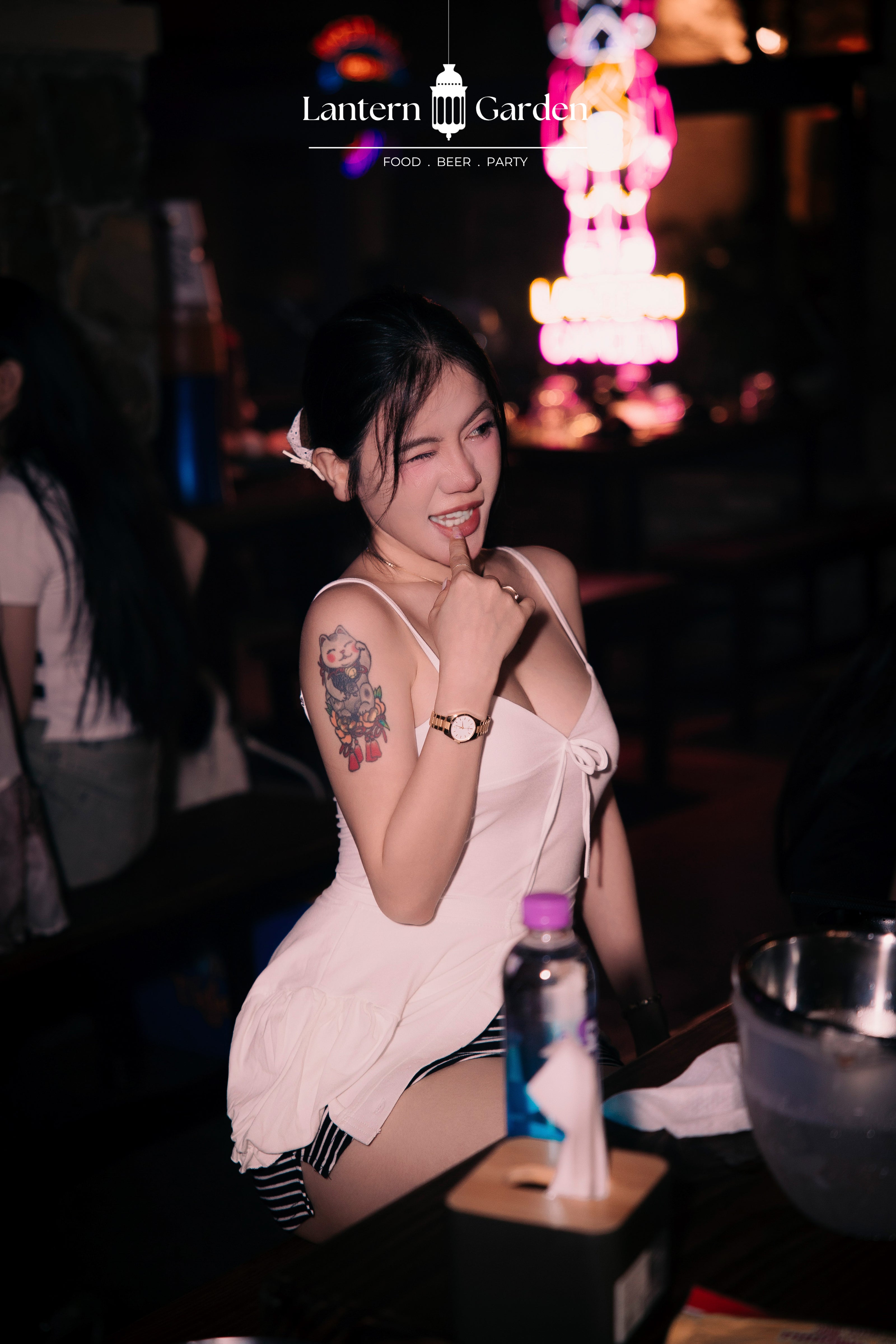 A woman posing in a dimly lit bar or club environment, wearing a white sleeveless top and striped shorts. A neon sign and the text 'Lantern Garden' are visible in the background.