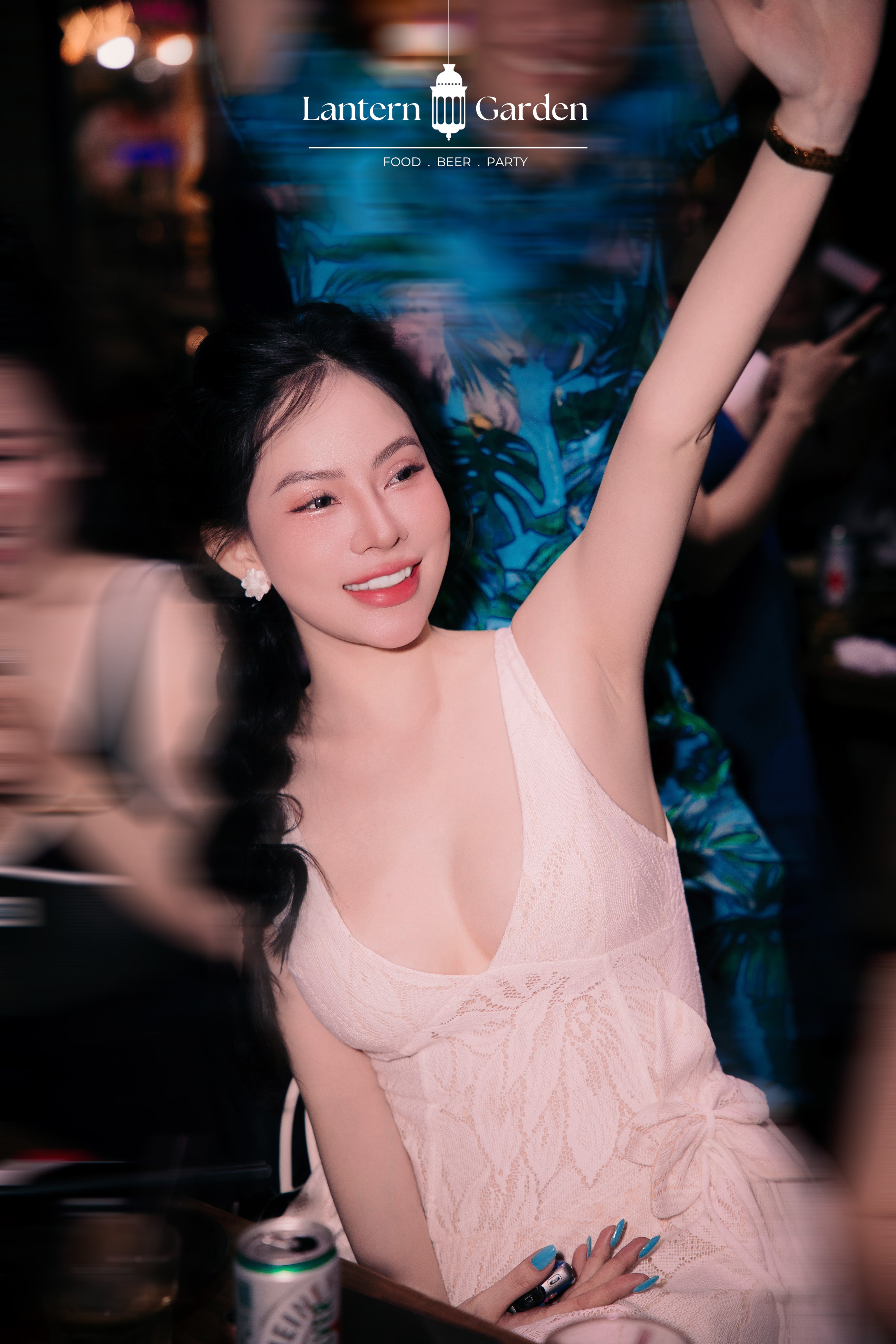A woman in a light pink lace dress smiling and raising her arm in a dimly lit social setting, with a visible 'Lantern Garden' logo at the top of the image.
