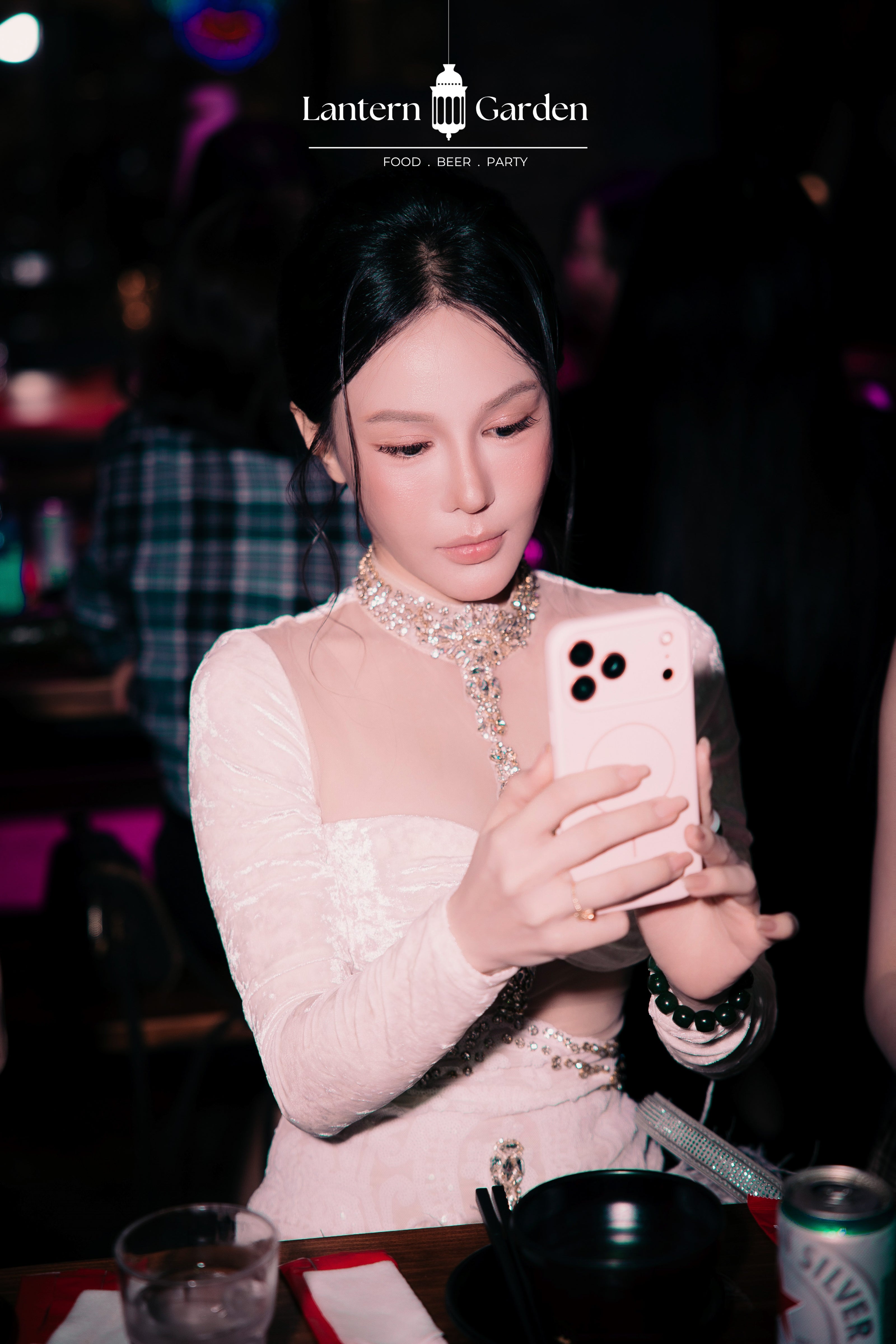 A woman in a light pink lace long-sleeve dress with a sparkling collar takes a mirror selfie in a dimly lit bar or restaurant setting. A white 'Lantern Garden' logo is visible at the top of the image.