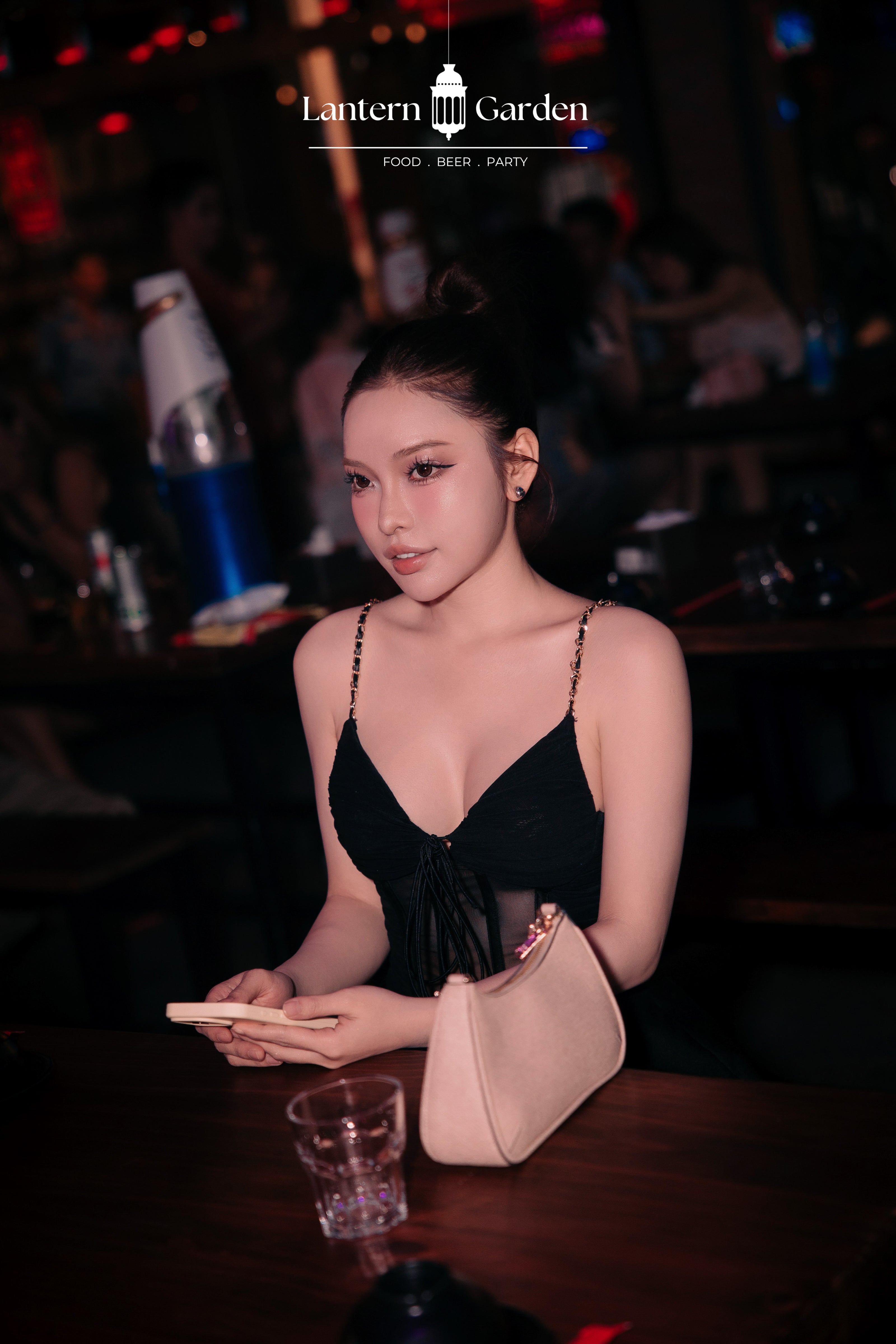 A woman in a black strappy top sits at a table in lantern garden nha trang, holding a light pink handbag and looking at her phone. The background is dark with warm red and blue ambient lighting, and the text 'Lantern Garden' is visible at the top.