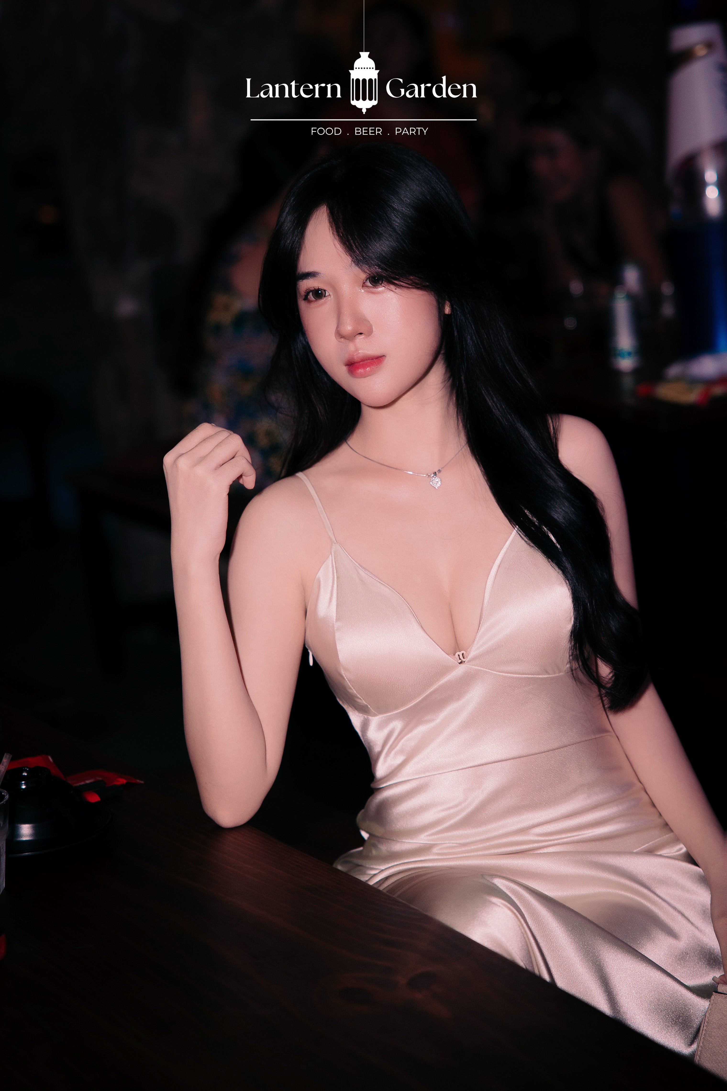 A woman with long black hair wearing a champagne satin spaghetti-strap dress, posing in a dimly lit bar or club setting. The 'Lantern Garden' logo is visible at the top of the image.