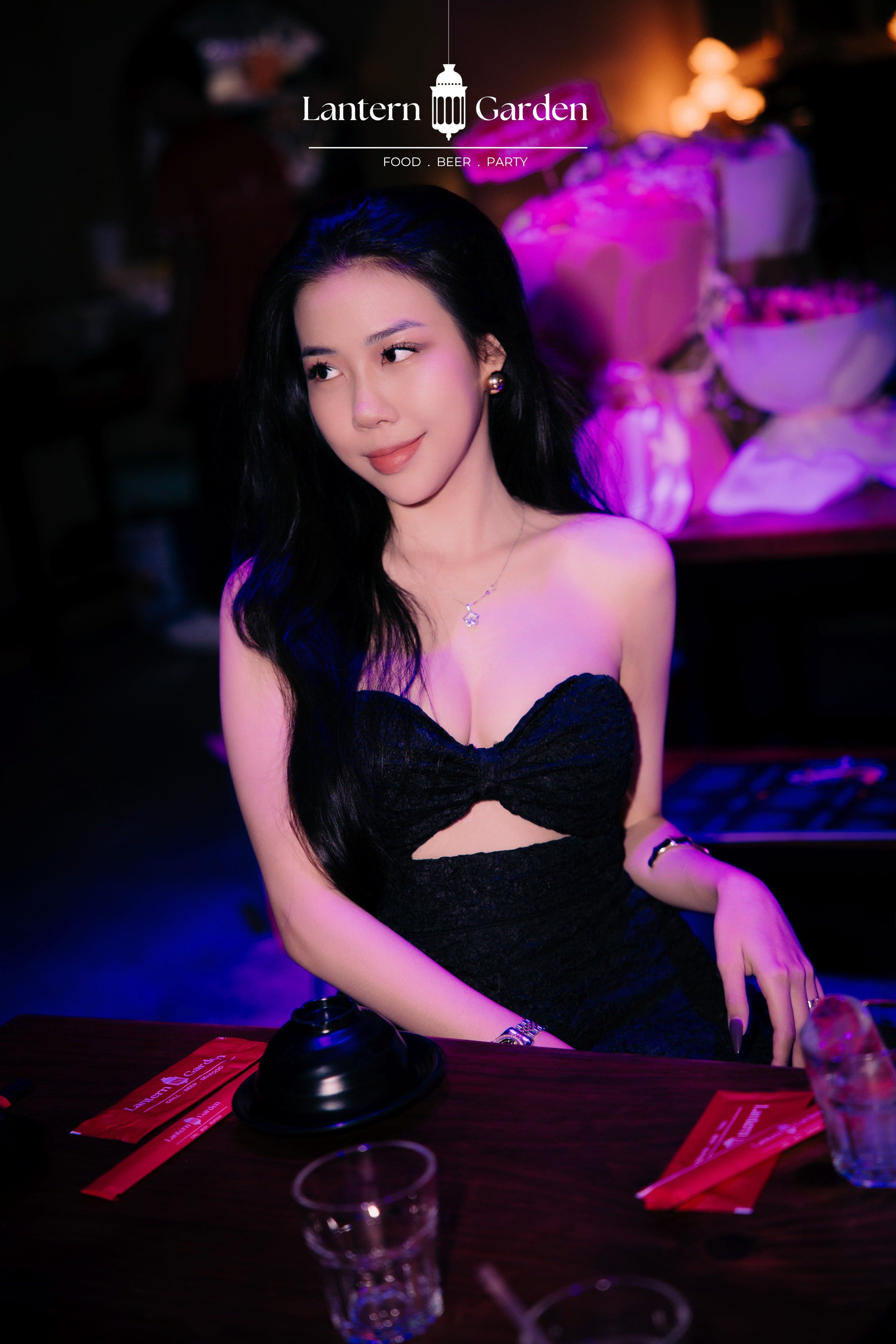 A woman in a black strapless sweetheart dress with a front cutout, seated at a bar in a dimly lit restaurant with purple and blue ambient lighting. The 'Lantern Garden' logo is visible at the top.