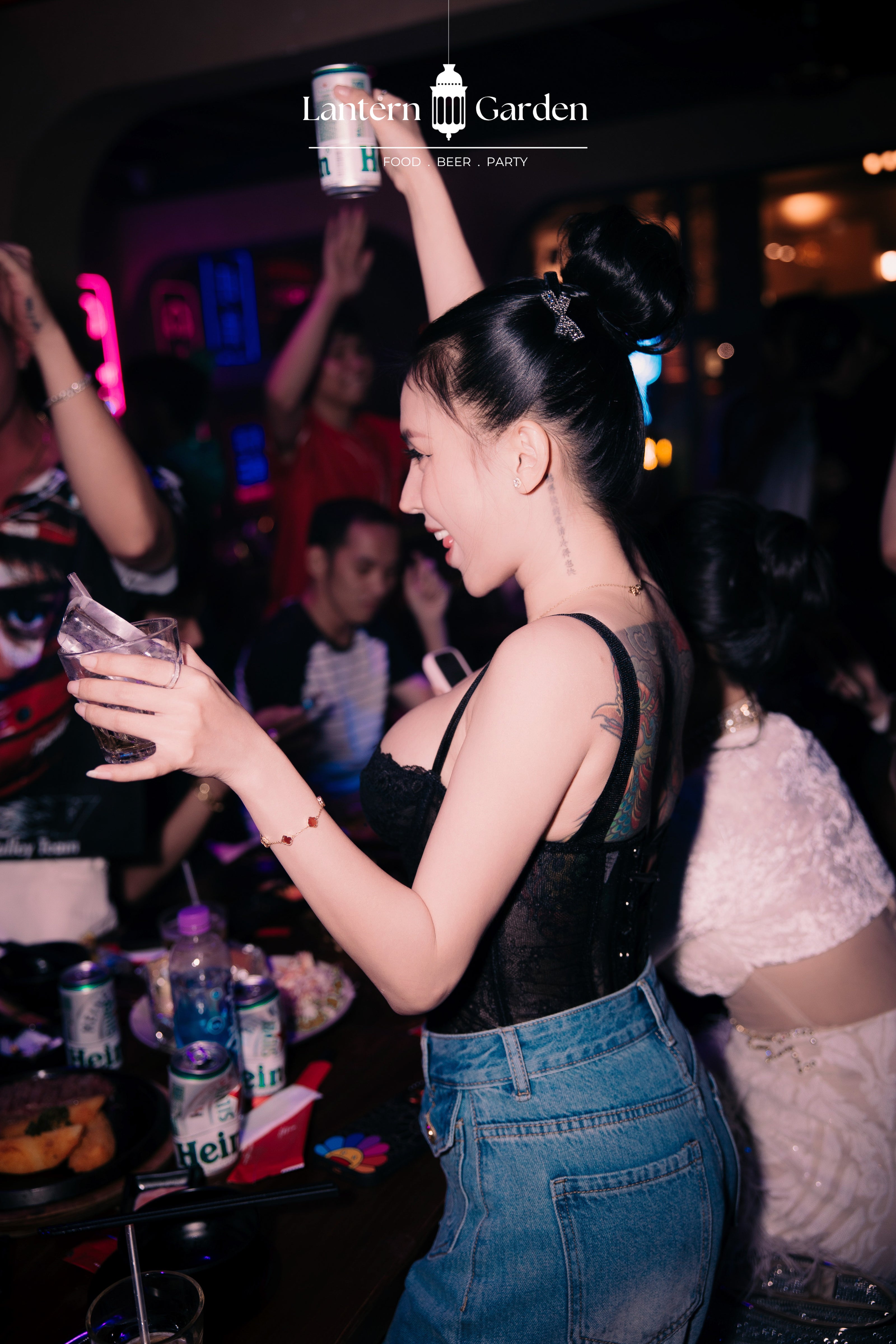 A woman in a black lace-trimmed camisole and blue denim jeans stands in a dimly lit nightclub or party setting, holding a drink. Other people and neon lighting are visible in the background, with a 'Lantern Garden Party' watermark at the top.
