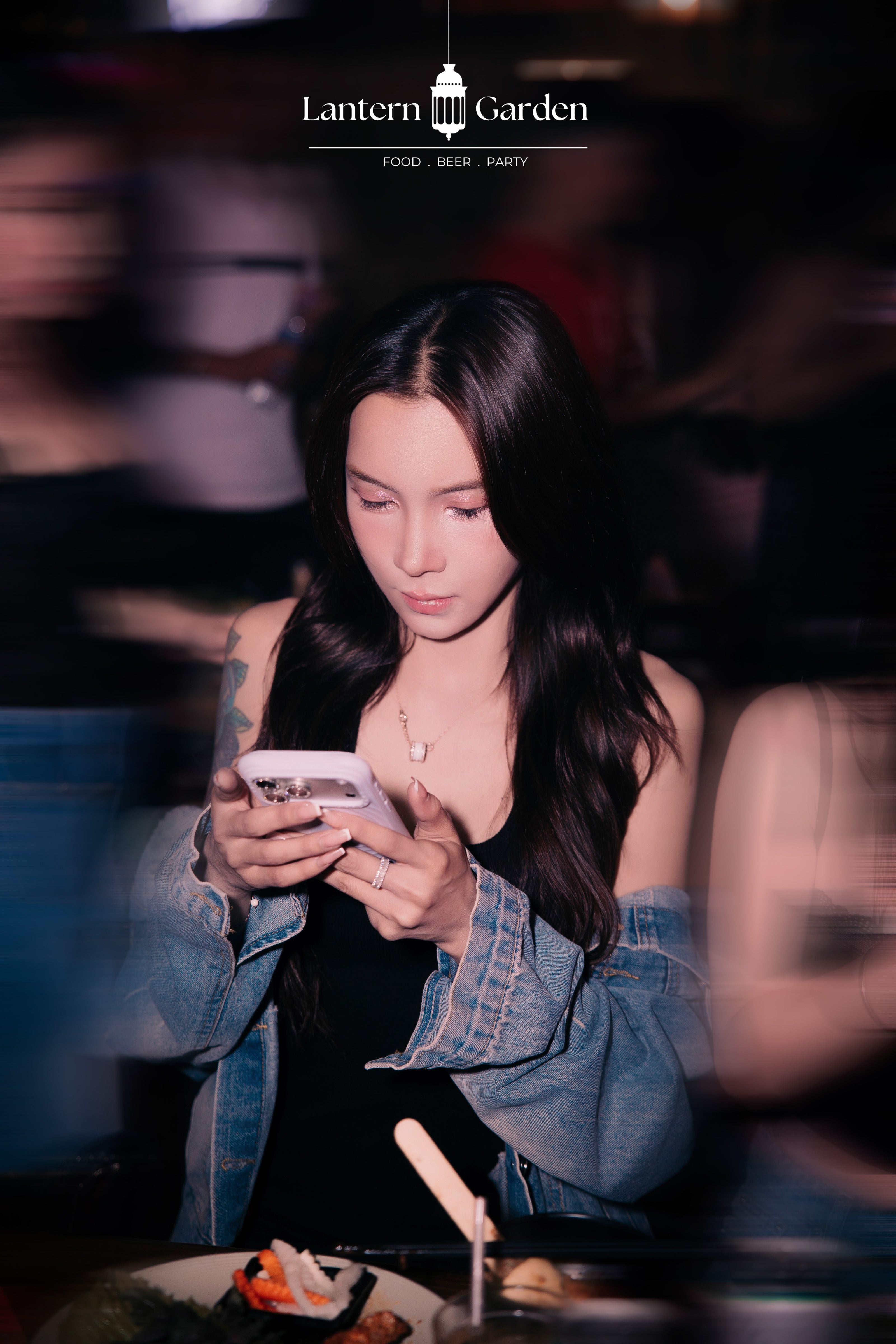 A woman with long dark hair looks down at a phone she is holding in a dimly lit social setting. She is wearing a light blue denim jacket over a black top.