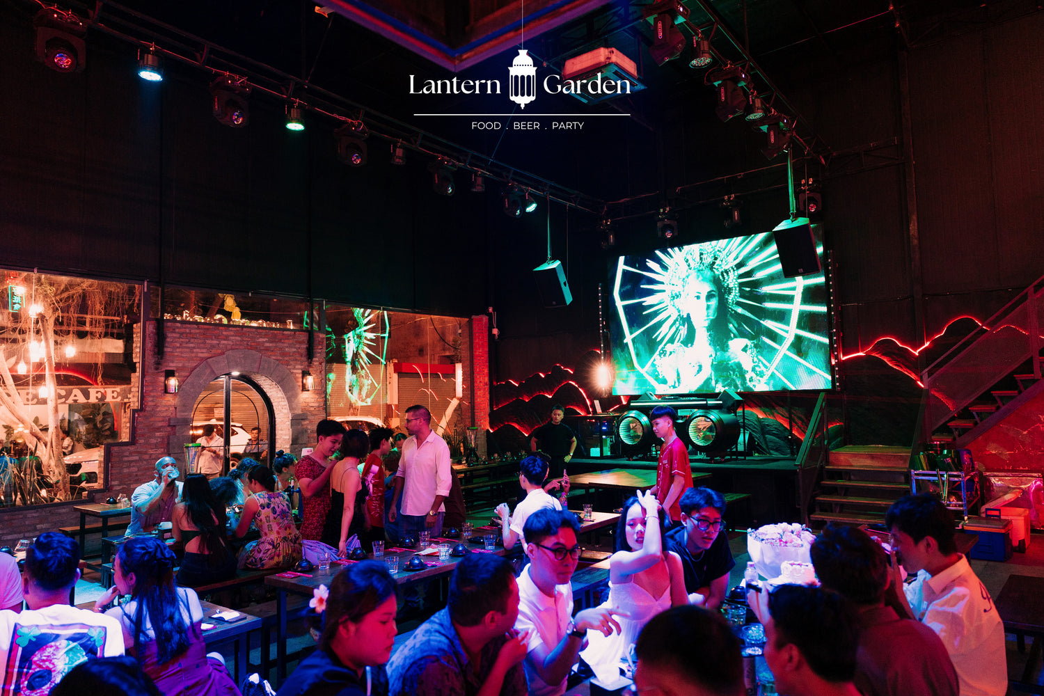 A crowded indoor event space with people dining and socializing under dramatic colored lighting. A large screen displays a green graphic, and the ceiling shows event lighting rigs and the text 'Lantern Garden' with 'FOOD | BEER | PARTY'.