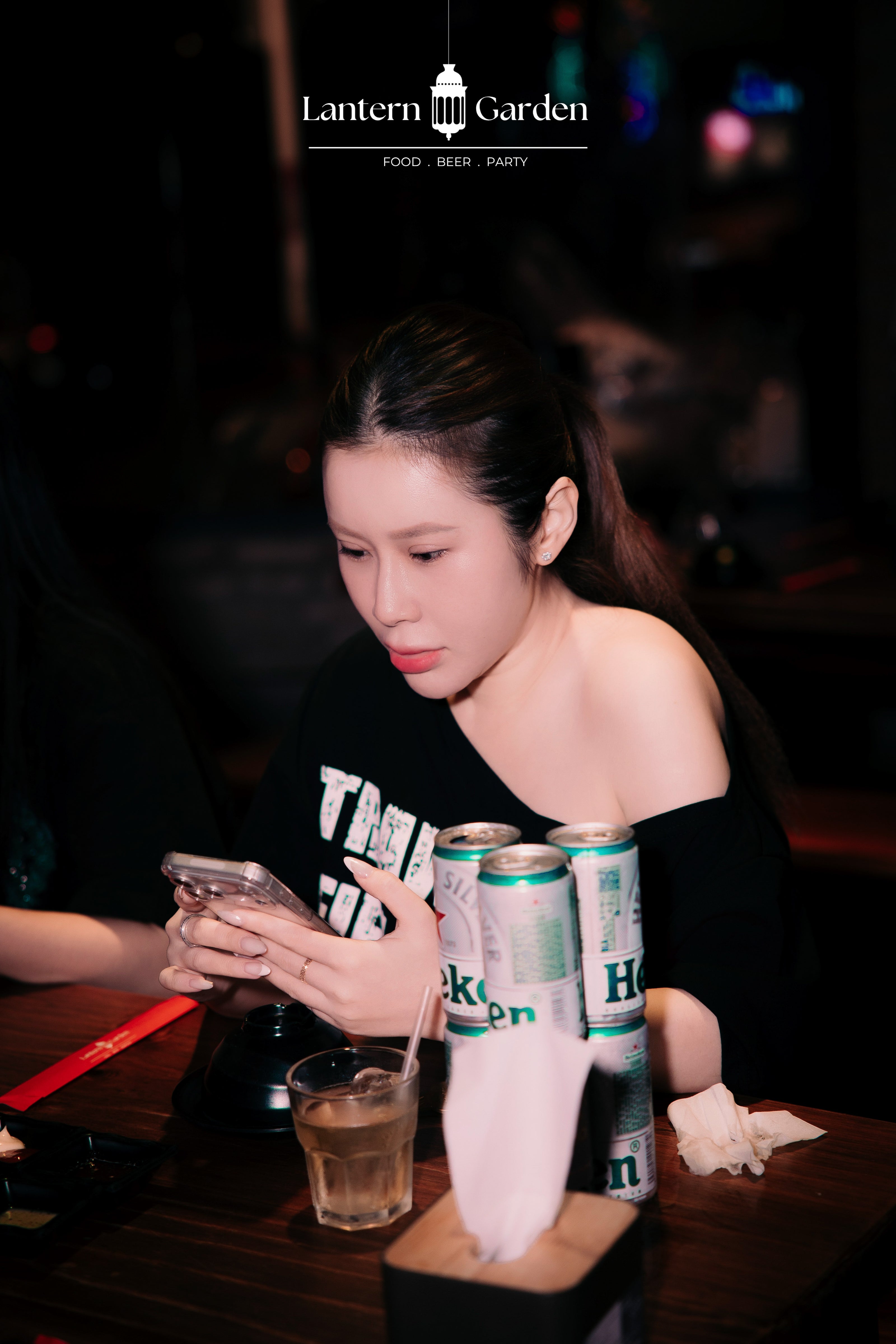 A woman in a black off-the-shoulder graphic shirt sits at a dimly lit bar table, looking down at her phone. Three cans of Heineken beer and a glass of beer are on the table in front of her.