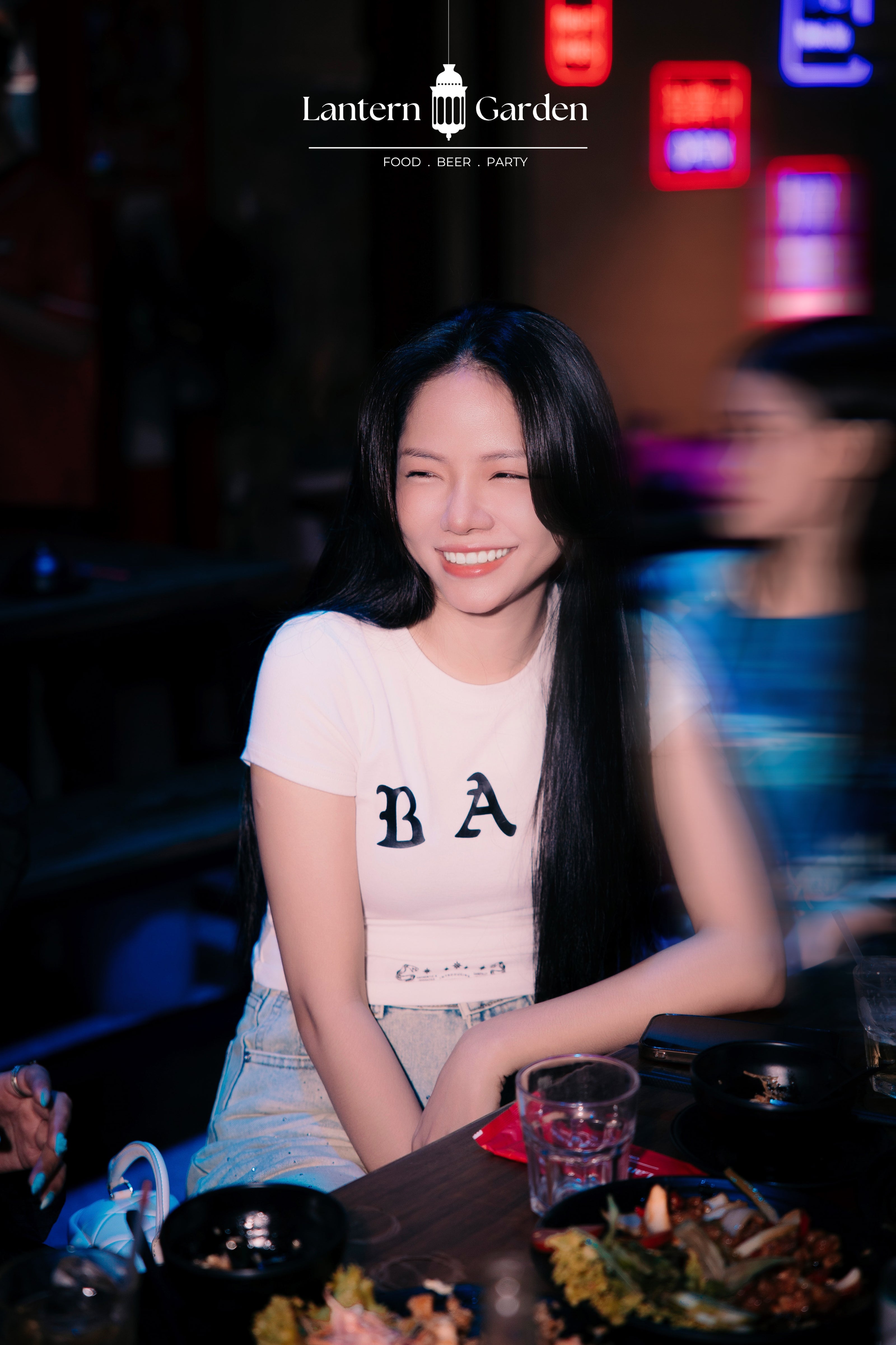 A woman smiling at a restaurant table wearing a white t-shirt with 'BA' printed on the front, with the 'Lantern Garden' logo at the top of the image.
