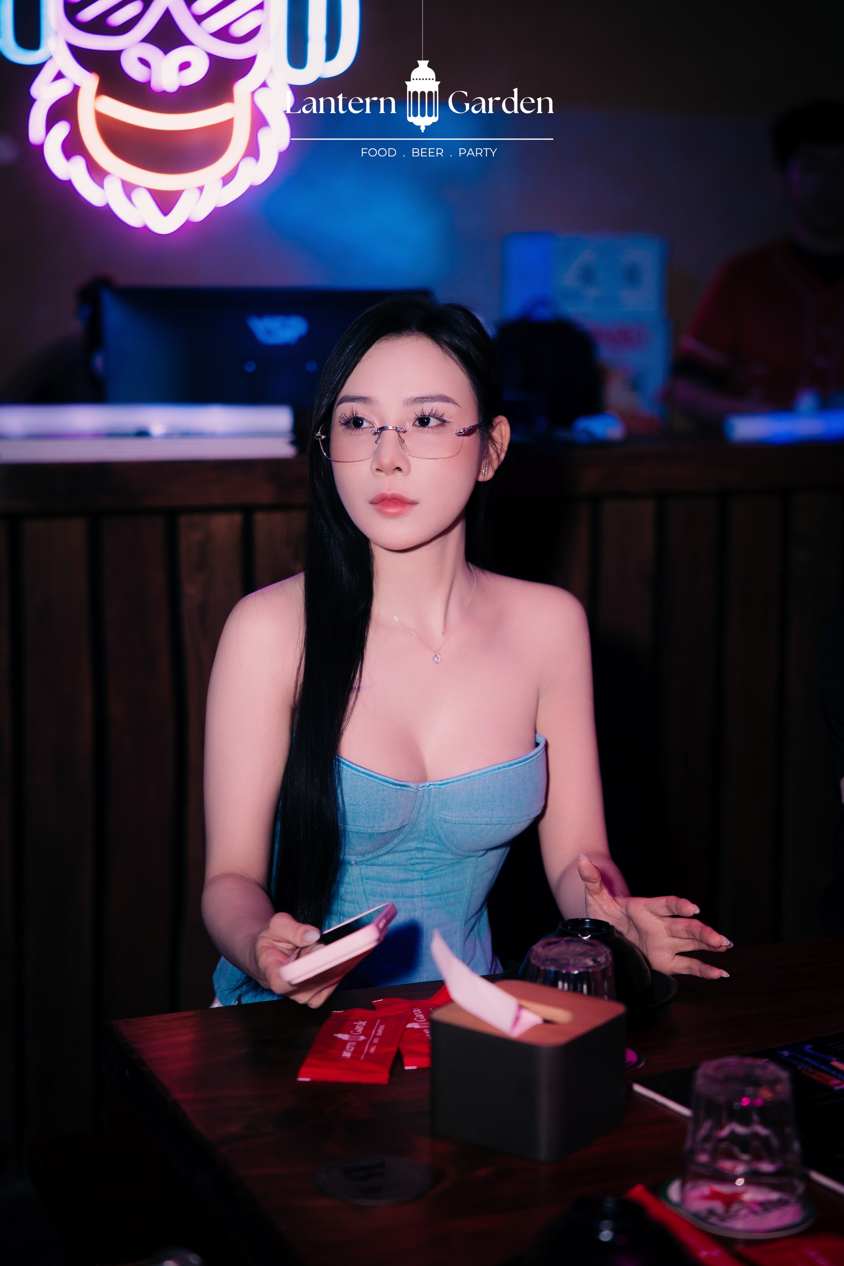 A woman with long black hair and glasses, wearing a light blue strapless top, sitting at a table in a dimly lit bar or restaurant. A neon sign is visible in the background, and the text 'Lantern Garden' appears at the top.