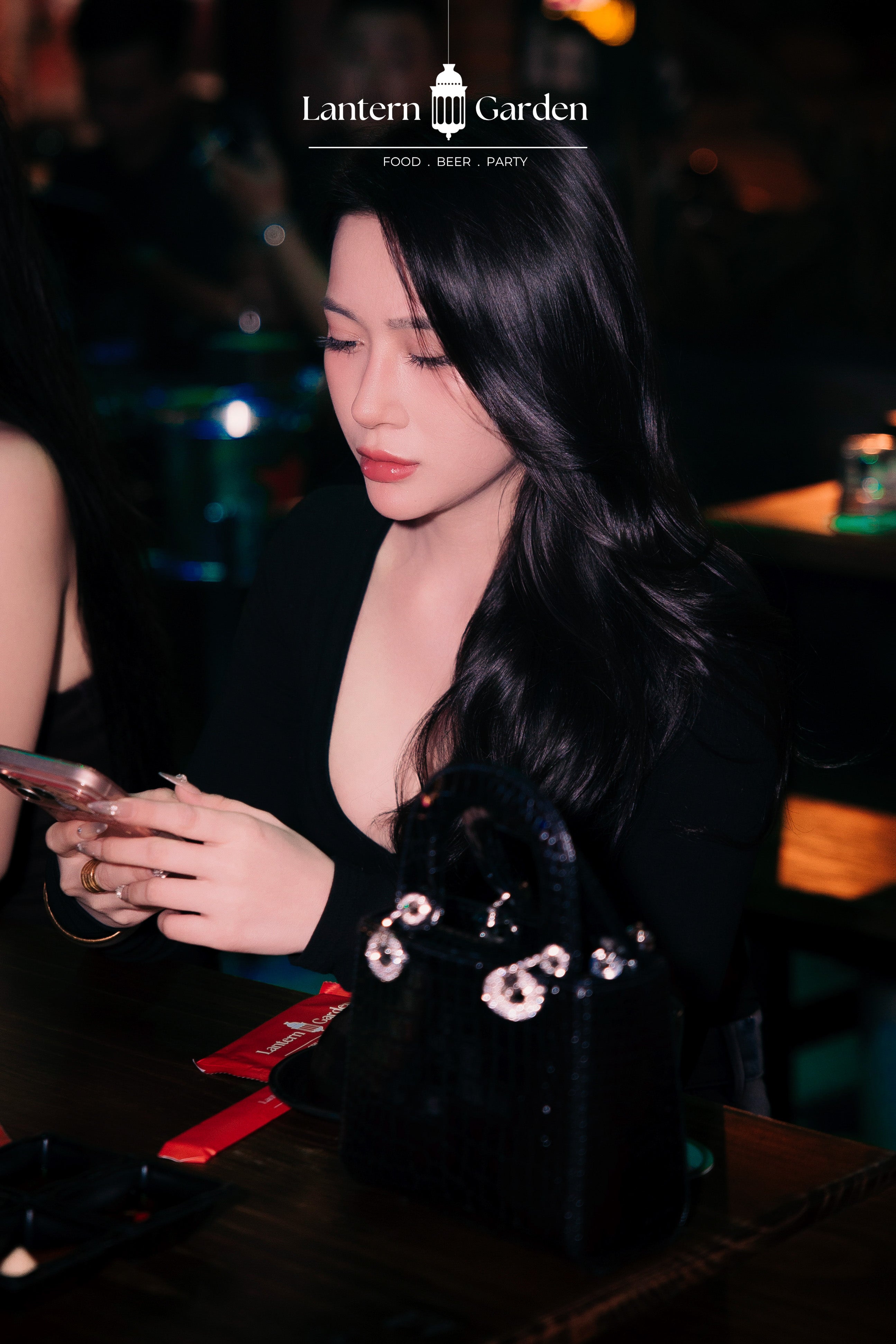 A woman in a black deep V-neck top looks down at a phone in a dimly lit social event setting. A black handbag with silver and white crystal embellishments sits on the table next to her.
