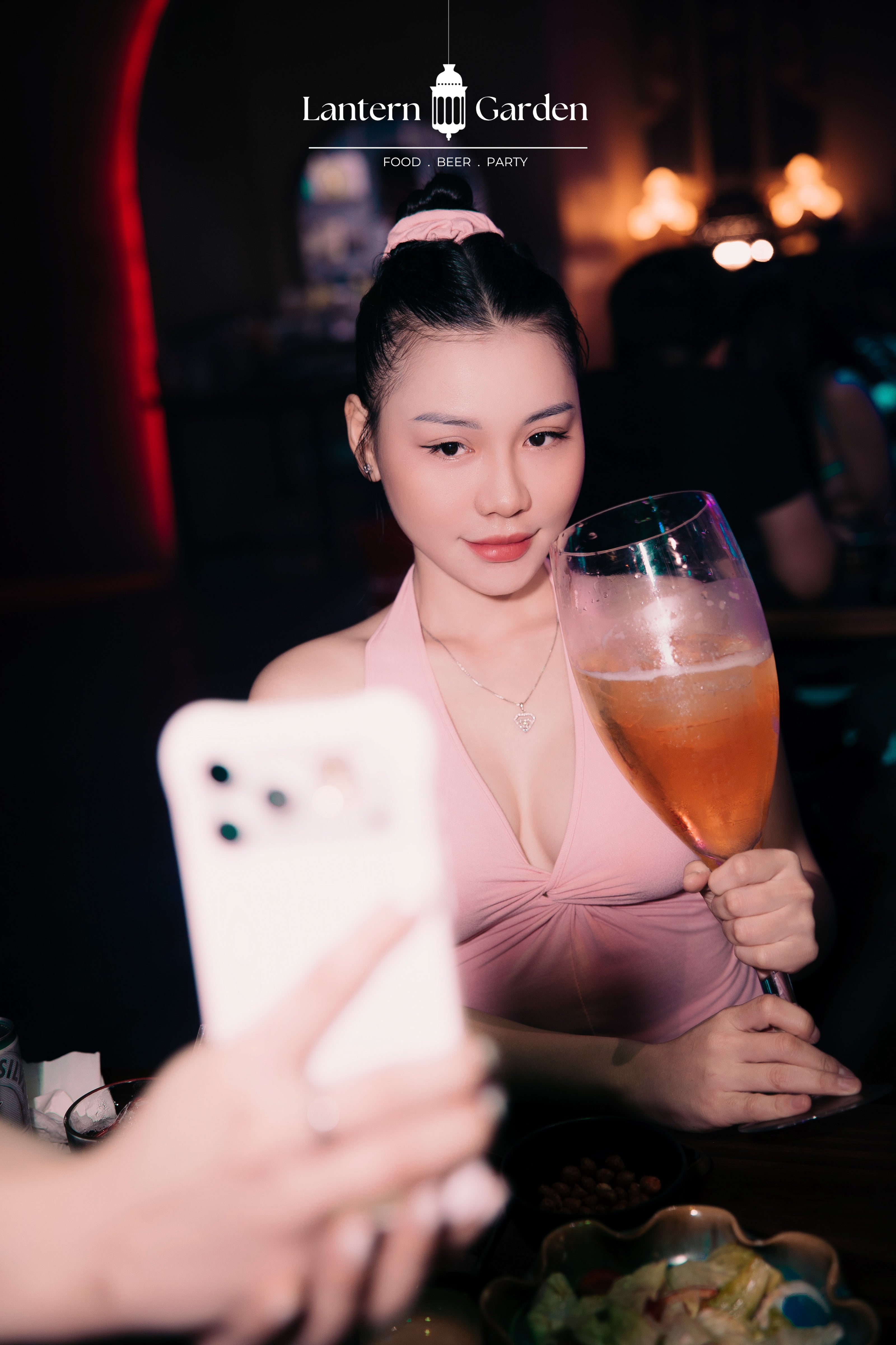 A woman in a pink halter dress holding a glass of amber-colored drink in a dimly lit bar or restaurant setting, with a phone in the foreground and the text 'Lantern Garden' at the top.
