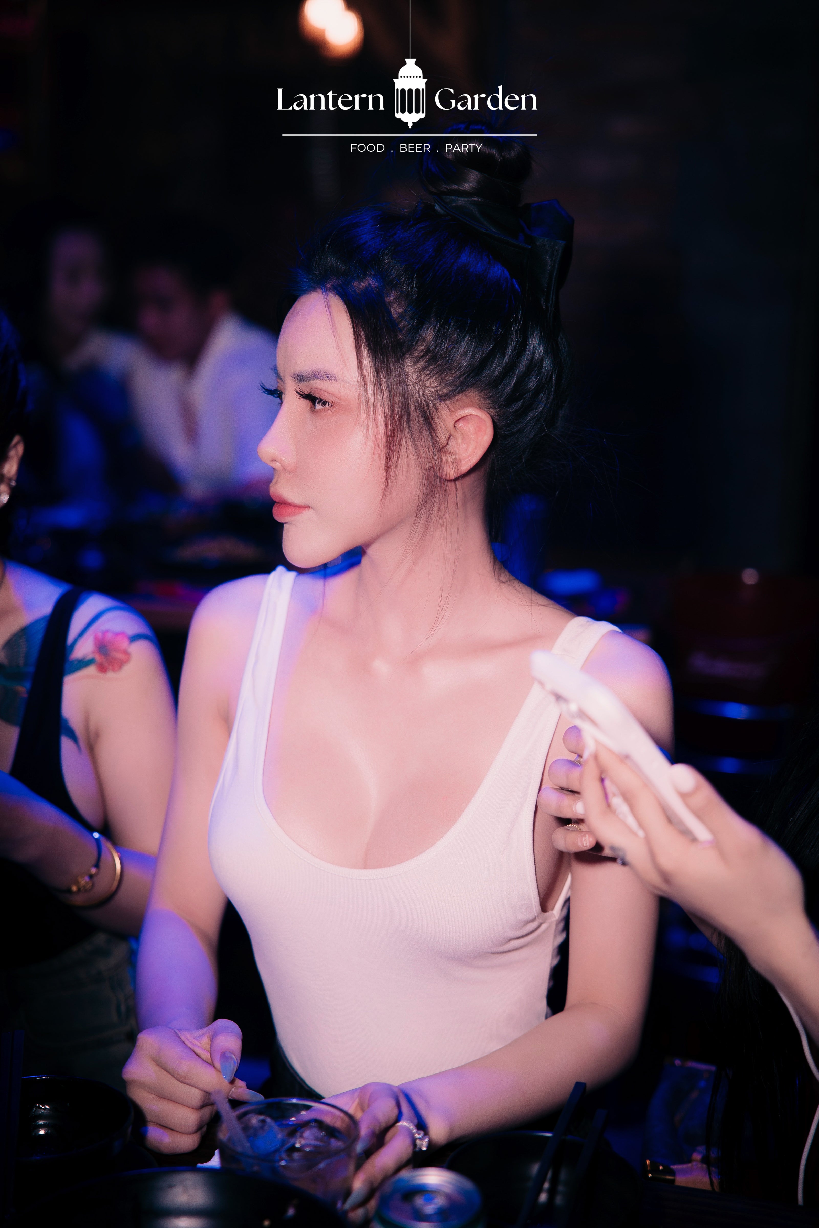 A woman in a tight-fitting white bodysuit at a dimly lit bar or club, with another person applying a bandage to her shoulder. The image has a 'Lantern Garden' logo at the top.