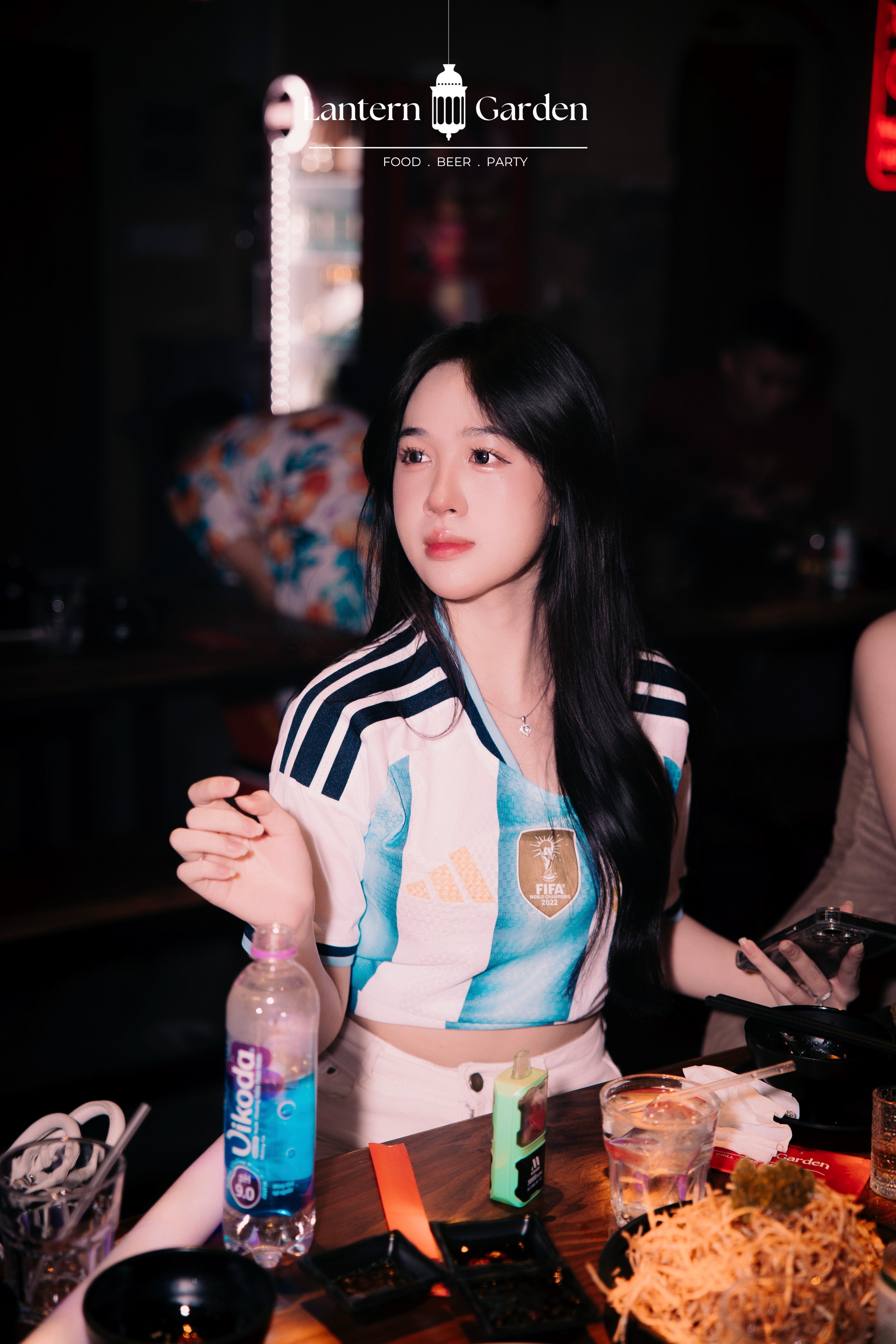 A woman wearing a white Argentina national football team Adidas jersey sits at a table in a dimly lit bar or restaurant, with drinks and food in the foreground. The text 'Lantern Garden' is visible at the top.