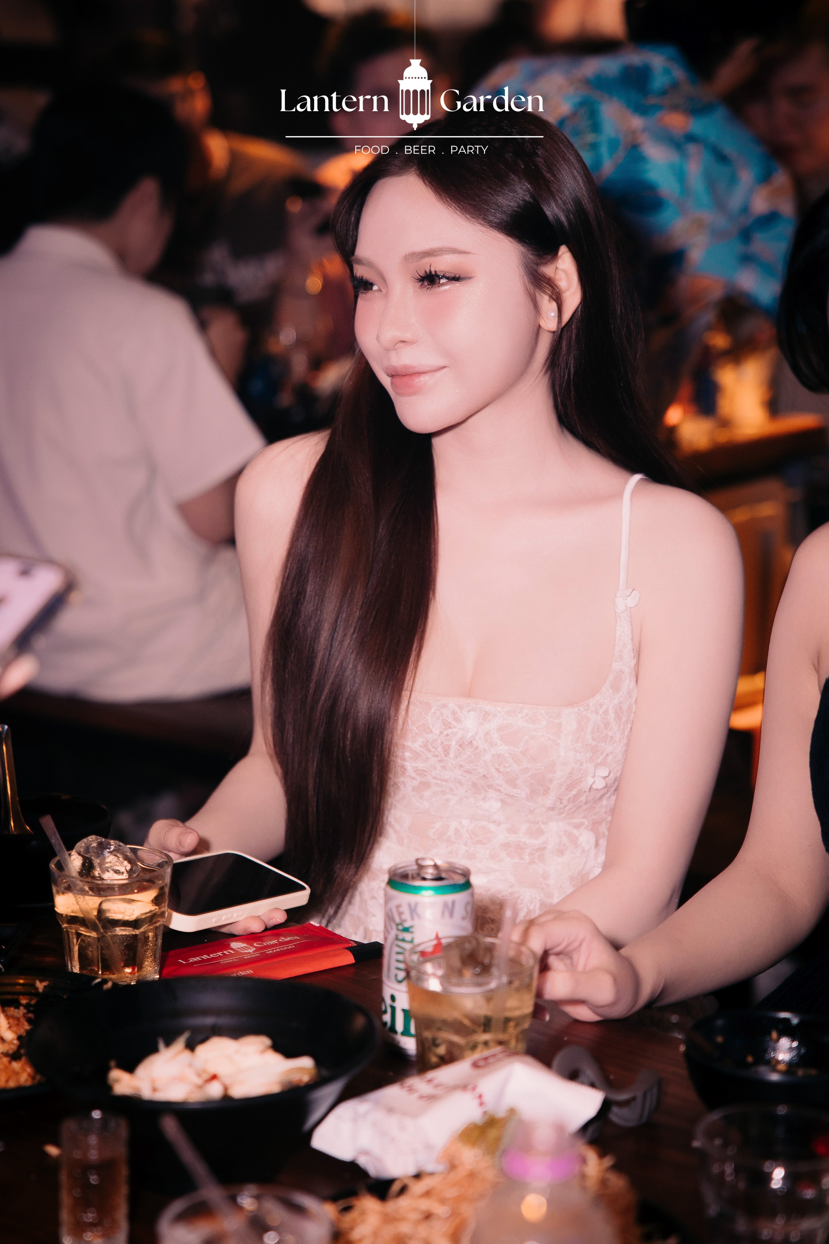 A woman with long dark hair wearing a white lace spaghetti-strap top sits at a restaurant table with drinks and food, under the Lantern Garden logo.