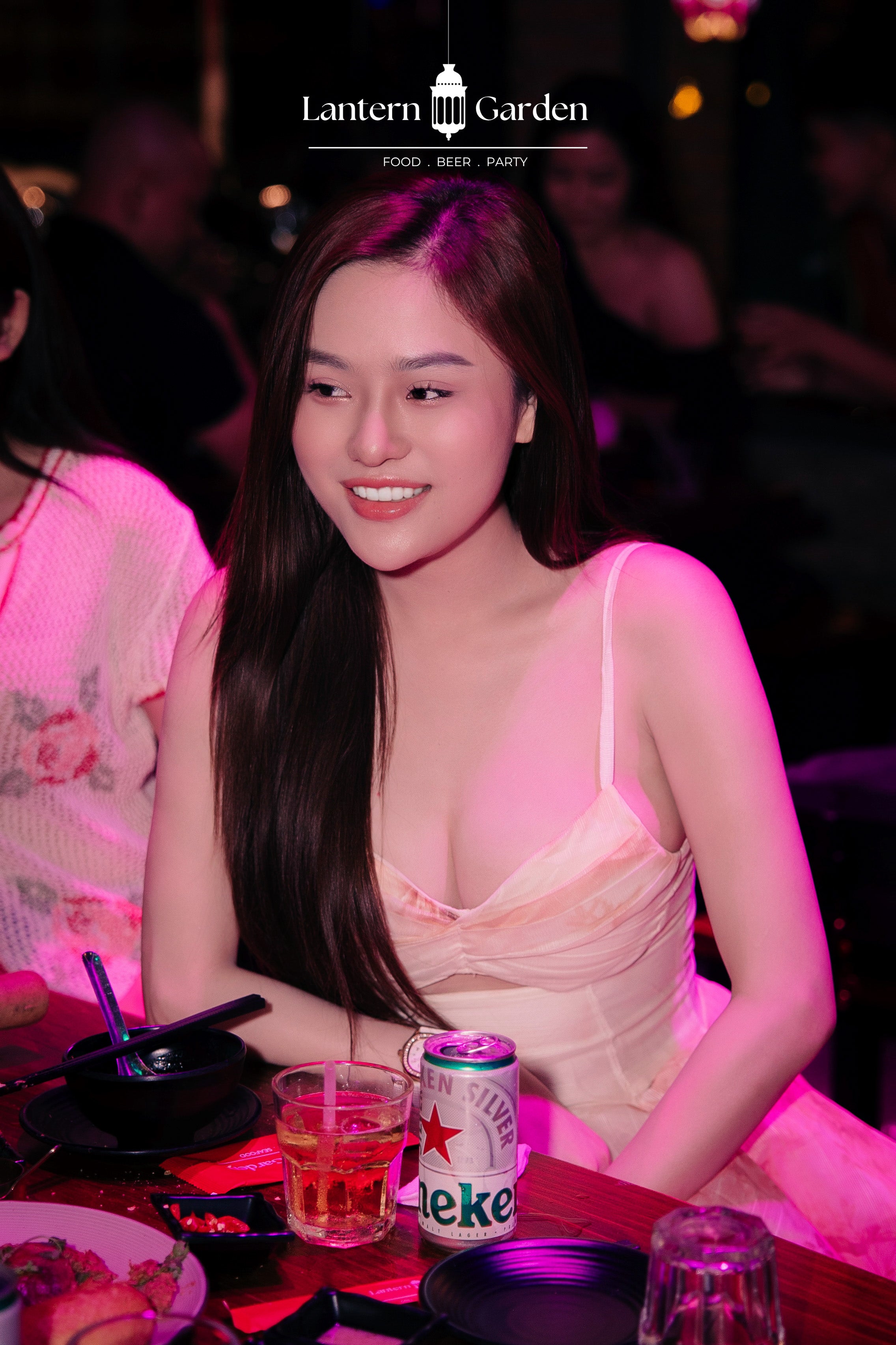 A woman with long dark hair smiling at a restaurant table, wearing a light pink spaghetti-strap dress under pink ambient lighting. A Heineken can and drinks are on the table in front of her.