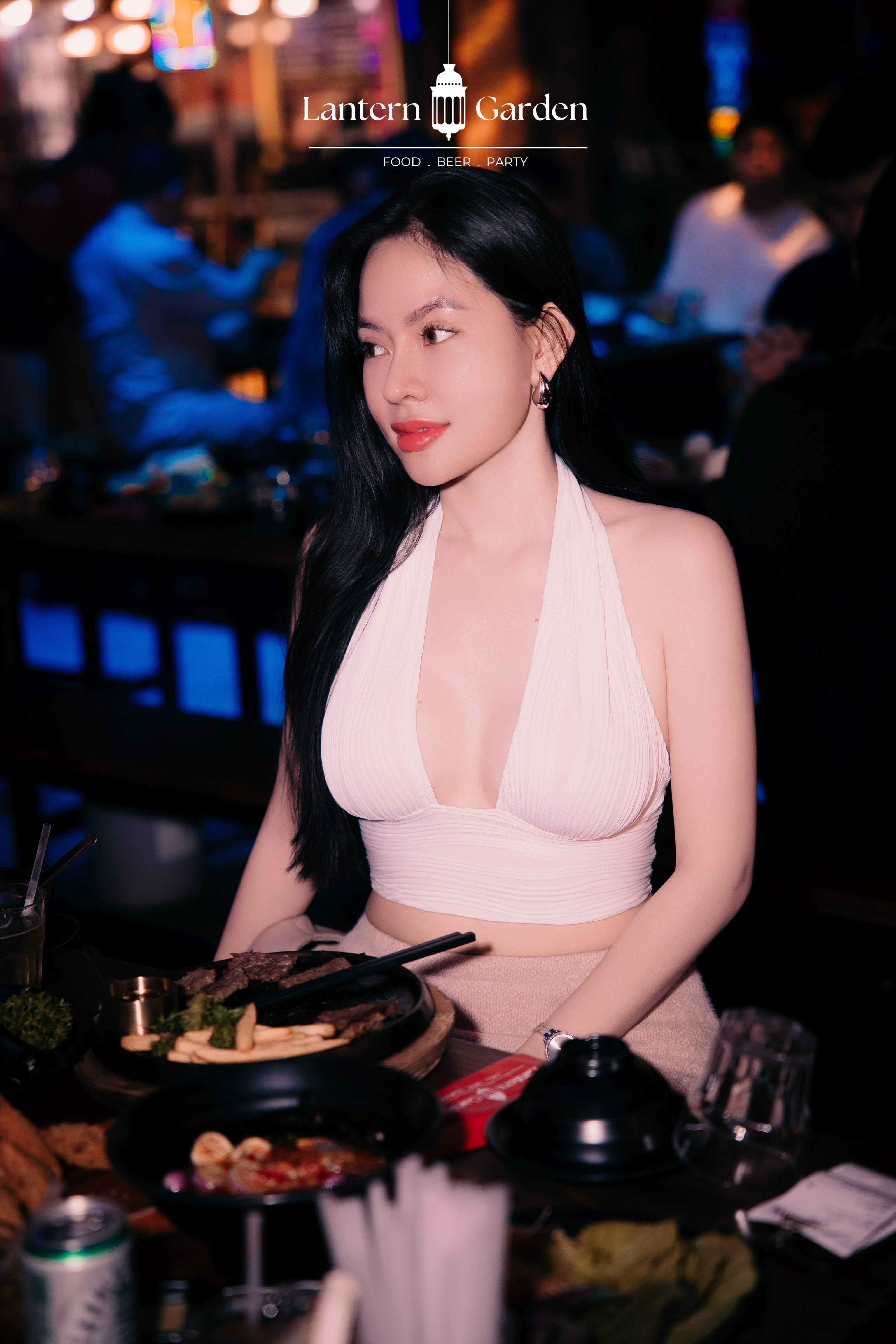 A woman seated at a restaurant table wearing a light pink halter crop top and matching skirt, with food and drinks in the foreground and a blurred bar or restaurant background. The text 'Lantern Garden' appears at the top.