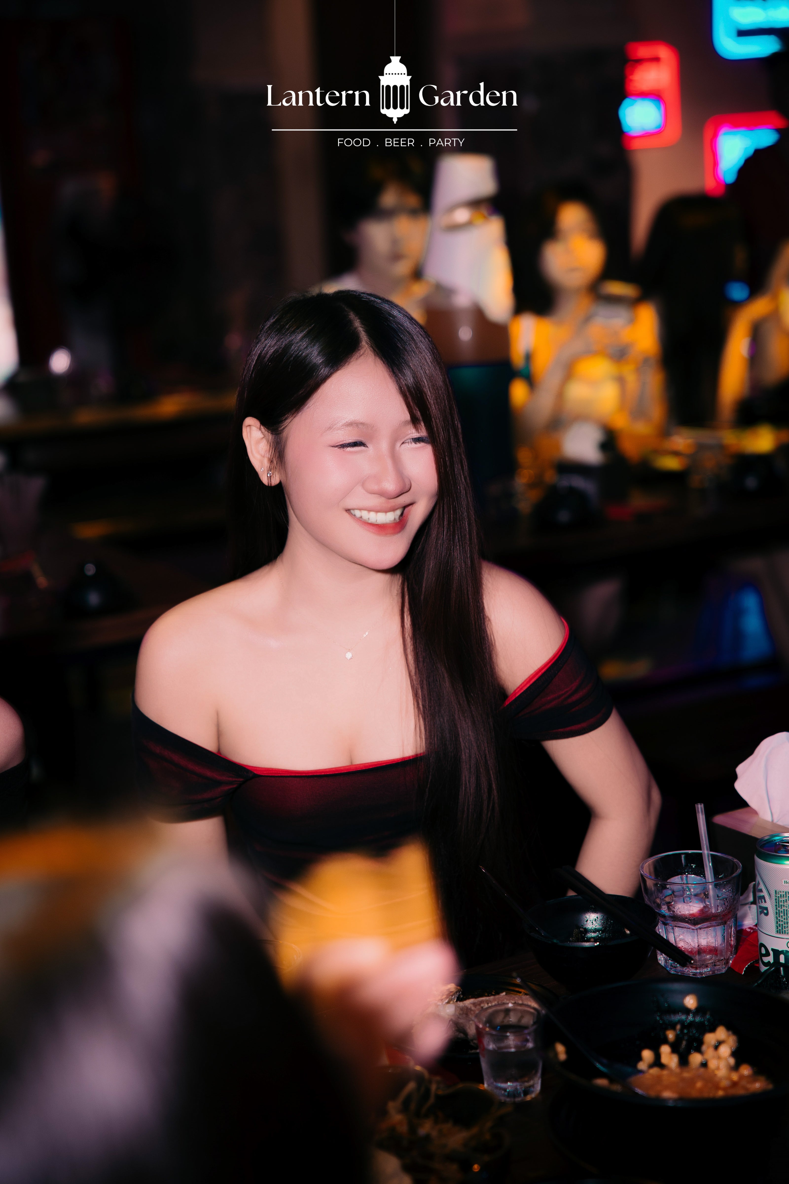 A smiling woman with long black hair is seated at a table in a dimly lit bar or restaurant, wearing a black off-the-shoulder top with red trim. The background is dark with warm ambient lighting and blurred neon signs.