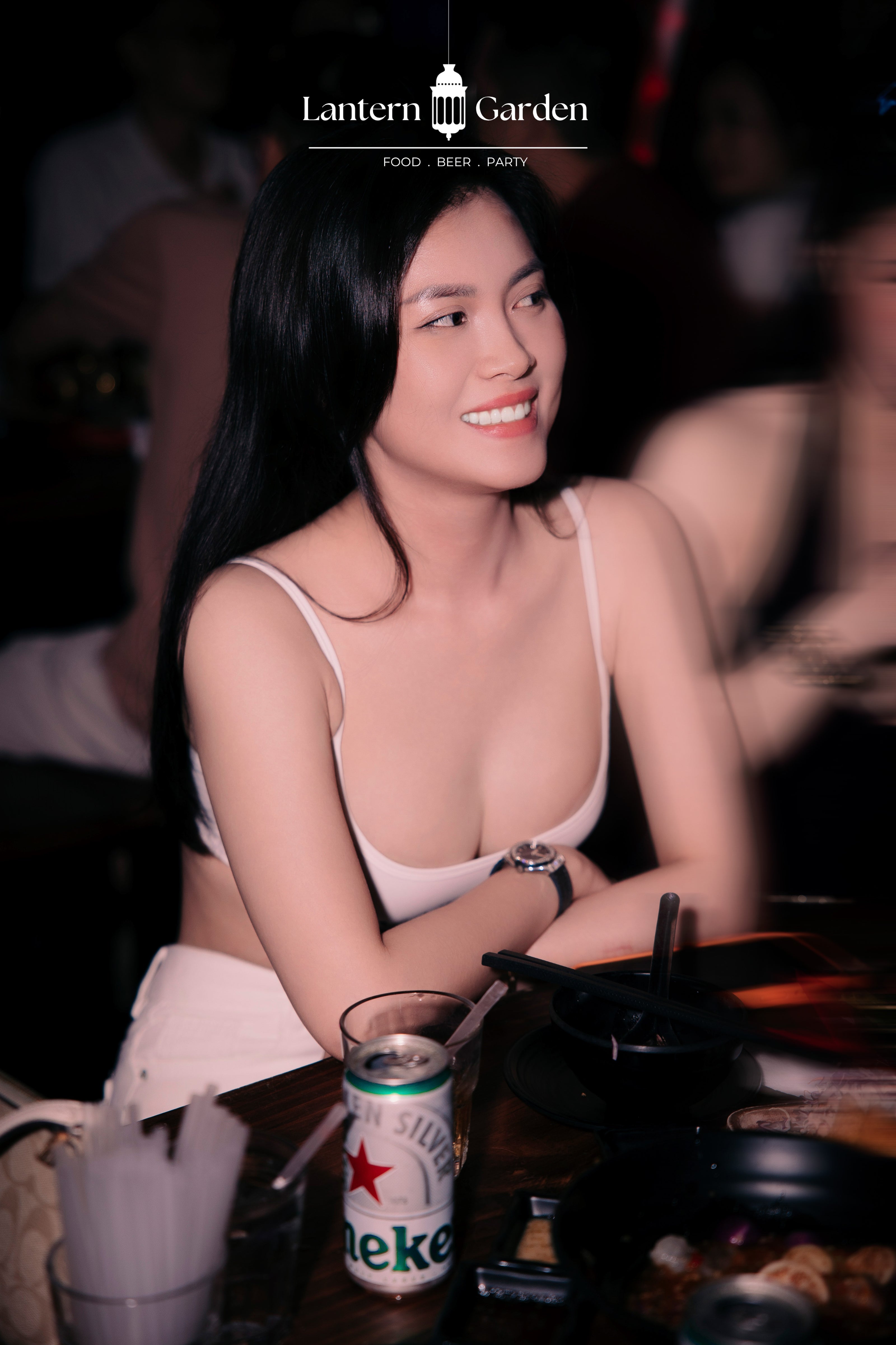 A woman with long black hair smiling at a restaurant table, wearing a light pink spaghetti-strap top. A Heineken beer can and a glass are in the foreground, with a 'Lantern Garden' logo overlay at the top.