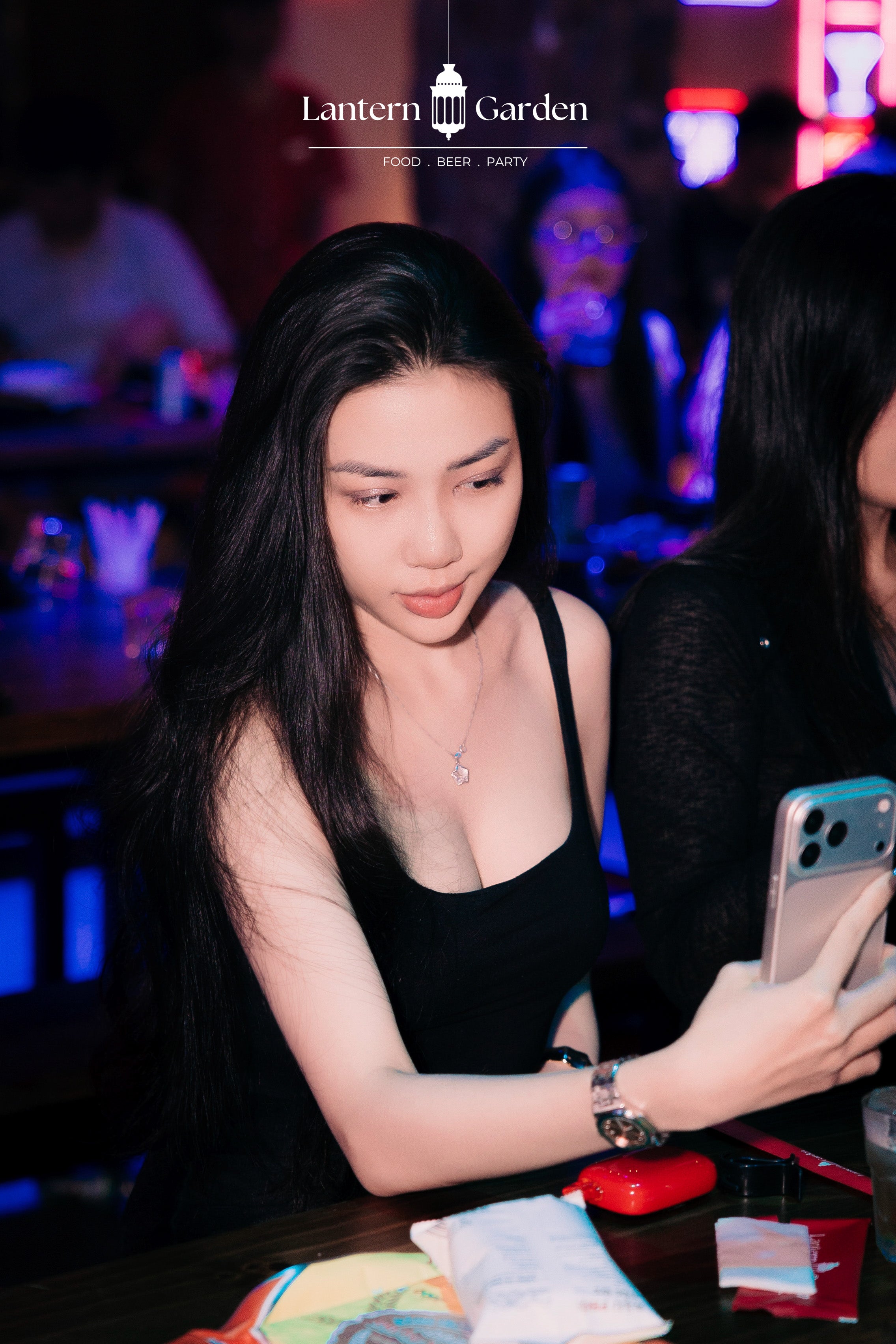 A woman in a black tank top holding a smartphone at a dimly lit bar or club, with the text 'Lantern Garden' visible at the top of the image.