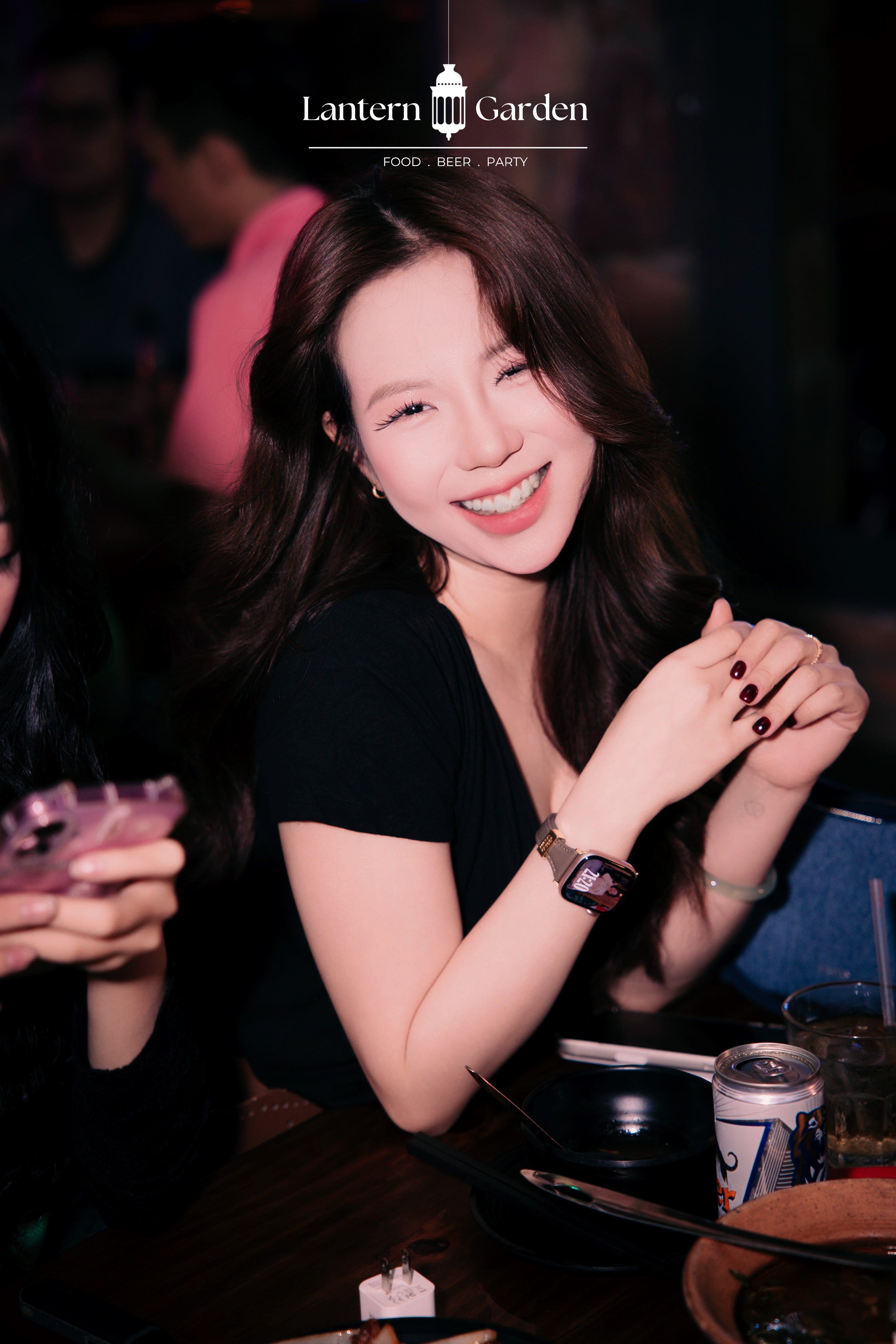 A smiling woman with long dark hair sits at a table in a dimly lit bar or restaurant, wearing a black top and a smartwatch. A can of beer and other dining items are visible on the table, and the 'Lantern Garden' logo is displayed at the top of the image.