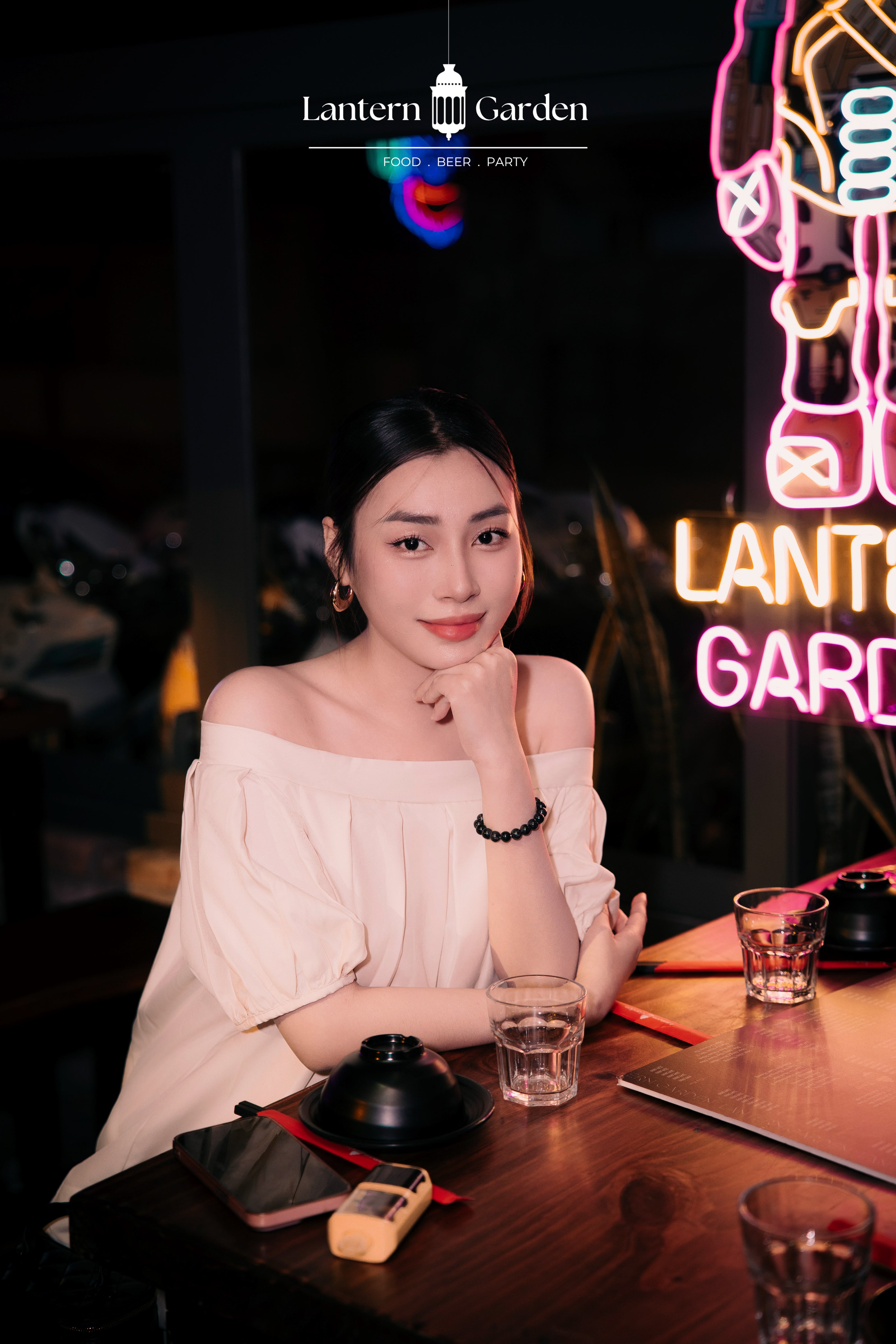 A woman wearing a cream off-the-shoulder blouse sits at a wooden table in a dimly lit restaurant or bar setting, with neon signs and glasses on the table. The brand name 'Lantern Garden' is visible at the top of the image.
