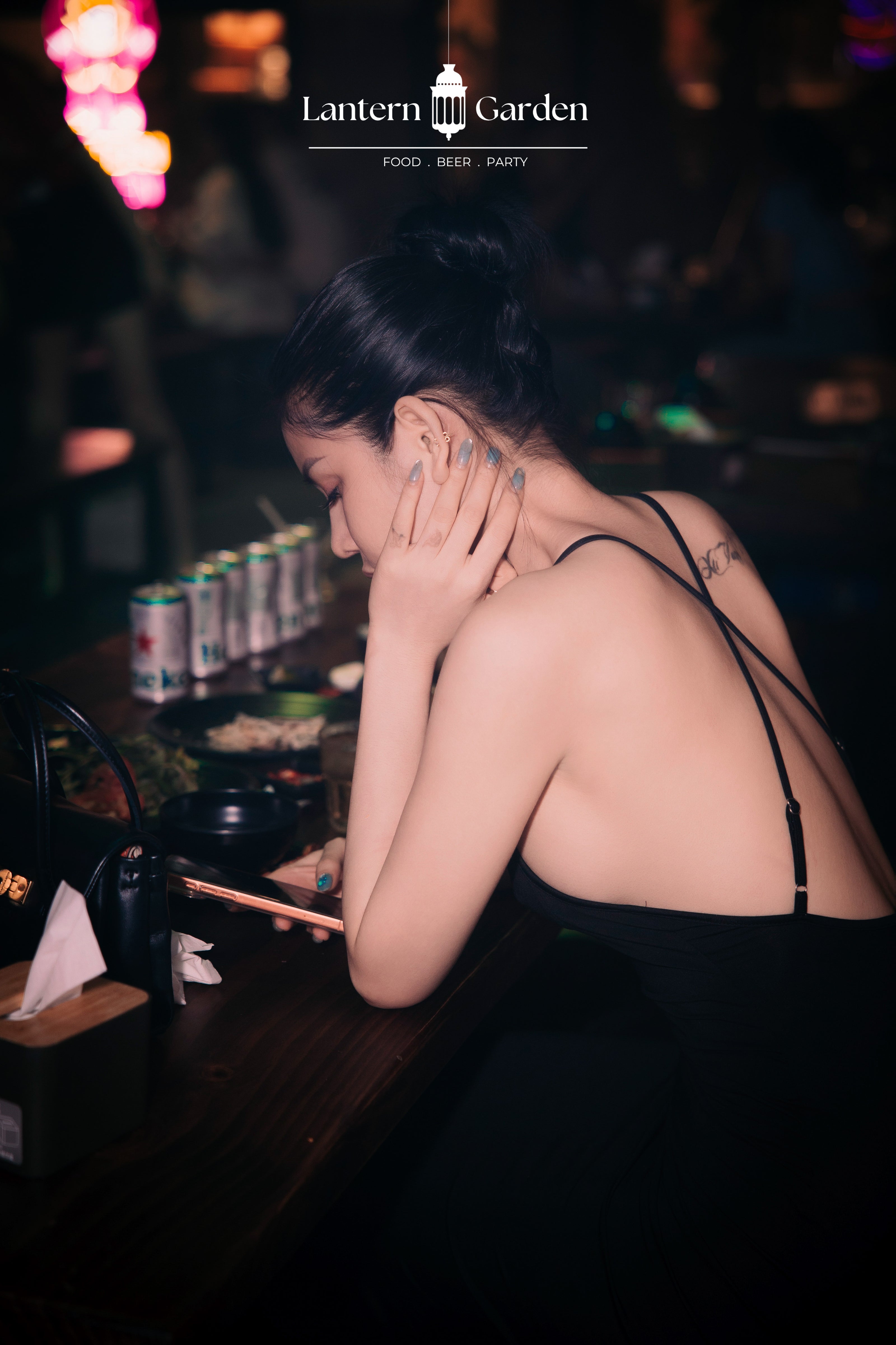 A woman in a black spaghetti-strap dress sits at a dimly lit bar, looking down with her hand near her face. The background features a dark, moody atmosphere with blurred lights and a row of beer cans on the counter. lantern garden nha trang