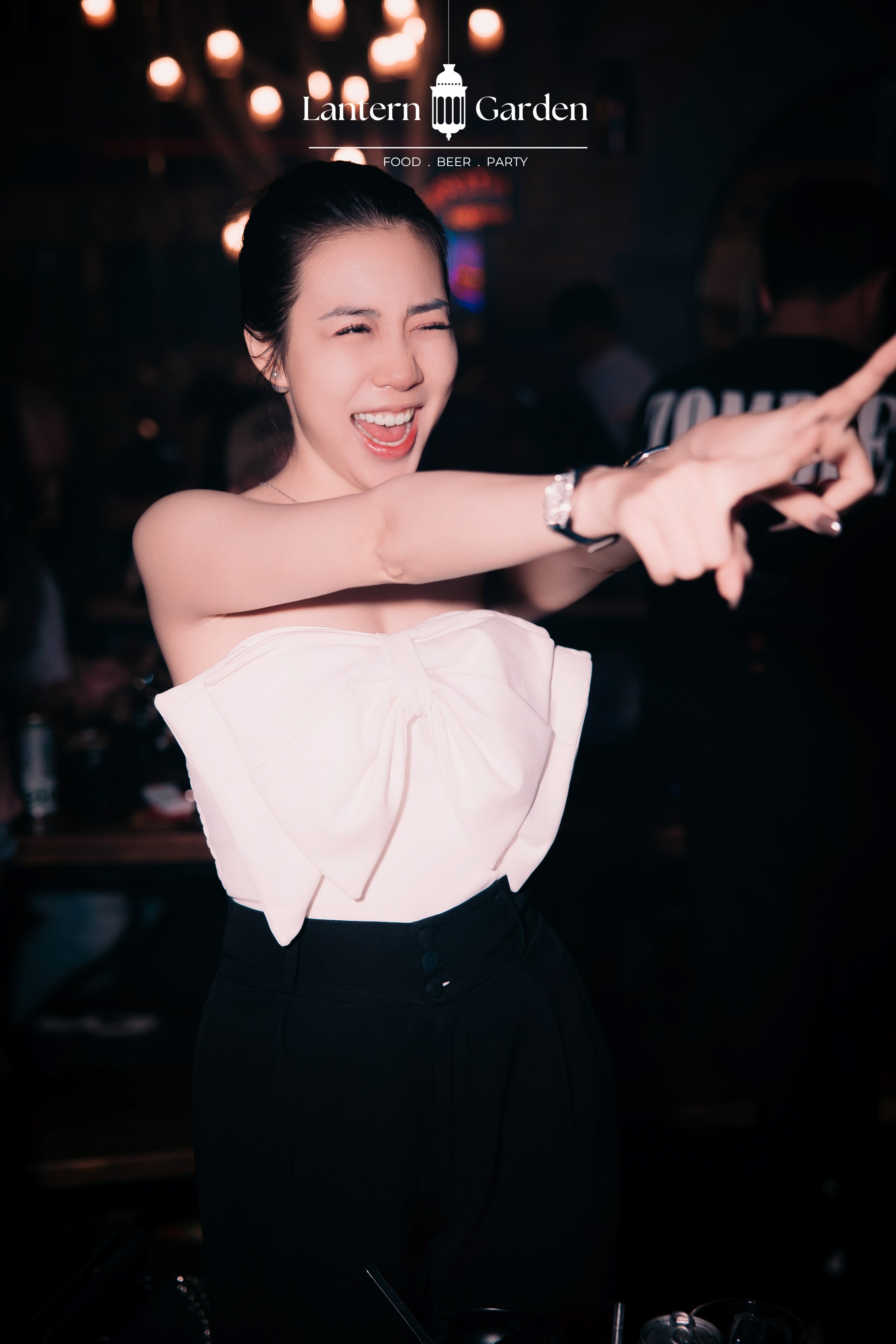 A woman in a black and white strapless top dancing in a dimly lit bar or club, with the 'Lantern Garden' logo visible at the top of the image.