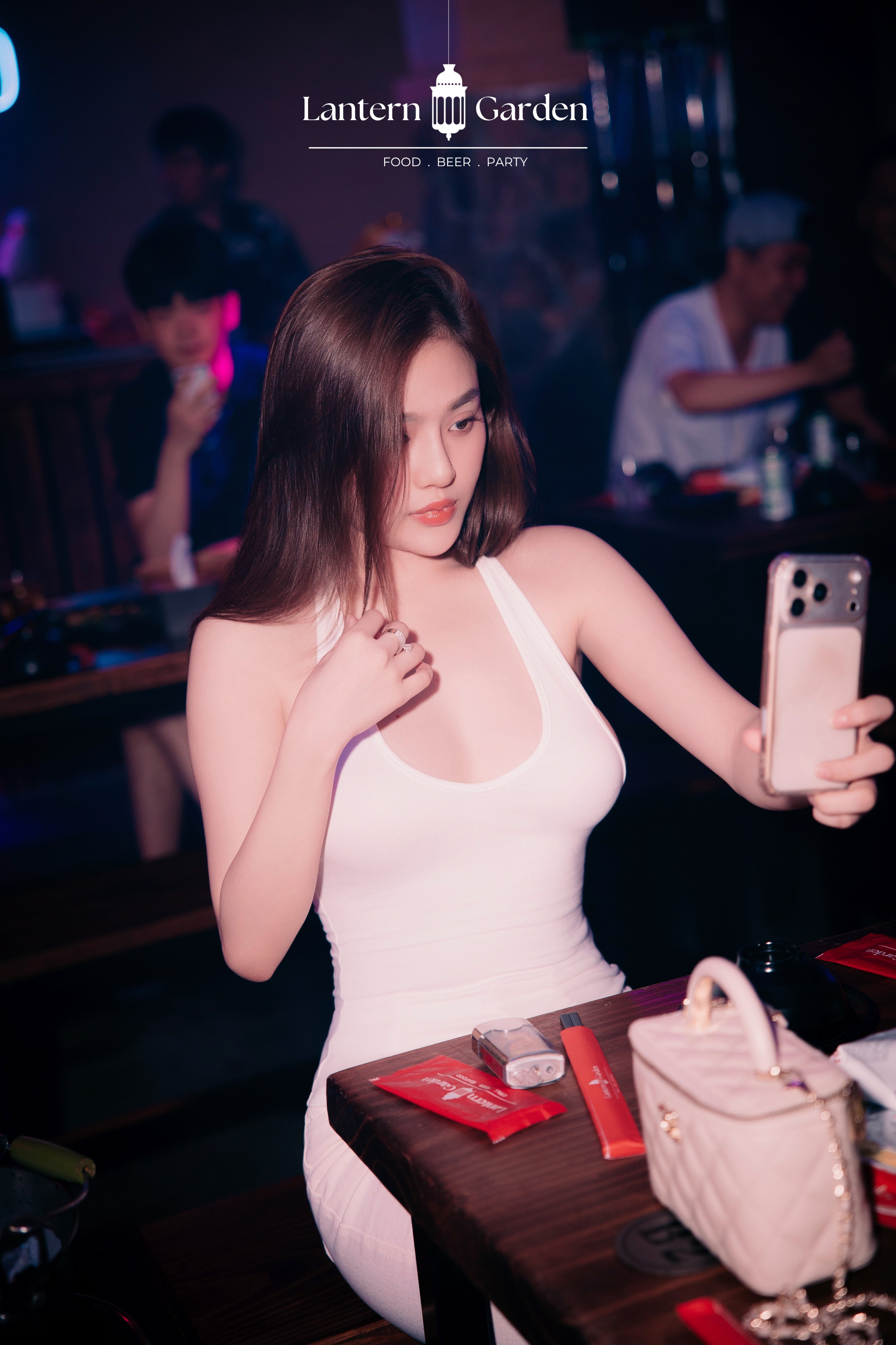 A woman in a tight white halter dress taking a mirror selfie in a dimly lit nightclub or bar, with a white handbag on the table in the foreground and the 'Lantern Garden' logo at the top.
