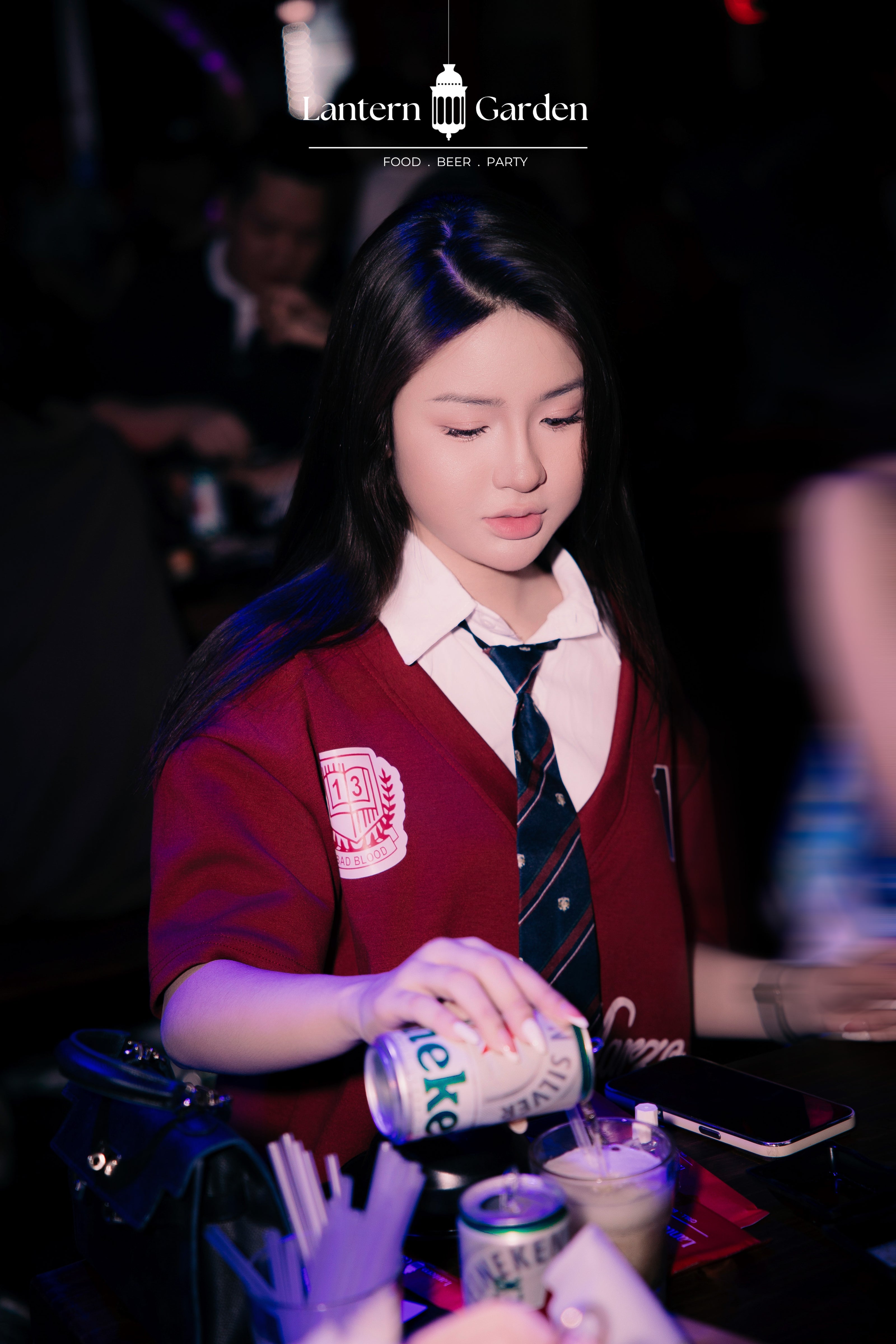 A woman in a school uniform-style red blazer, white collared shirt, and striped tie pours a can of beer at a dimly lit bar. The Lantern Garden logo is visible at the top, and the scene is photographed in low light with a blurred background.