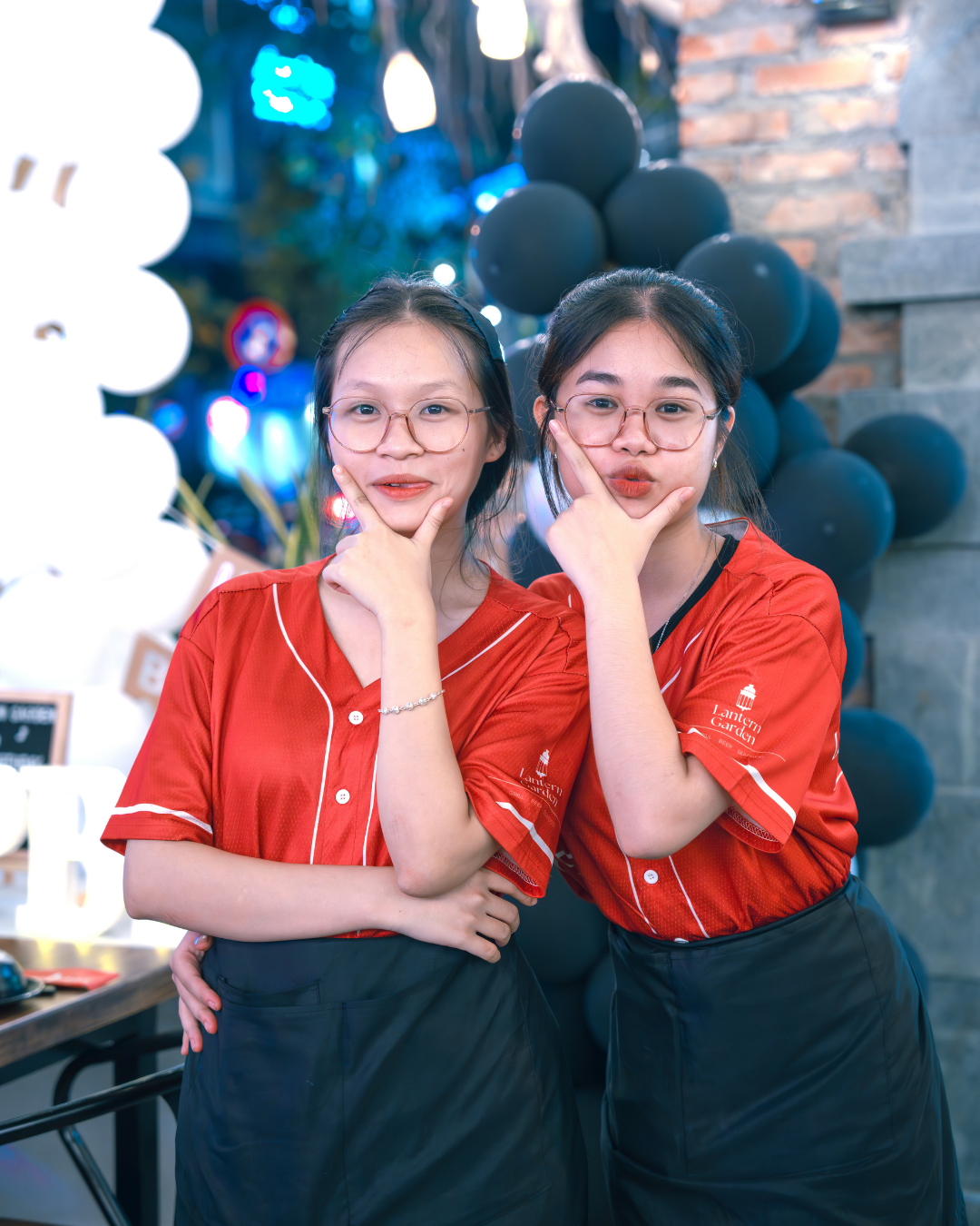 Two women in red shirts posing together with balloons and a tree with lantern in the background.